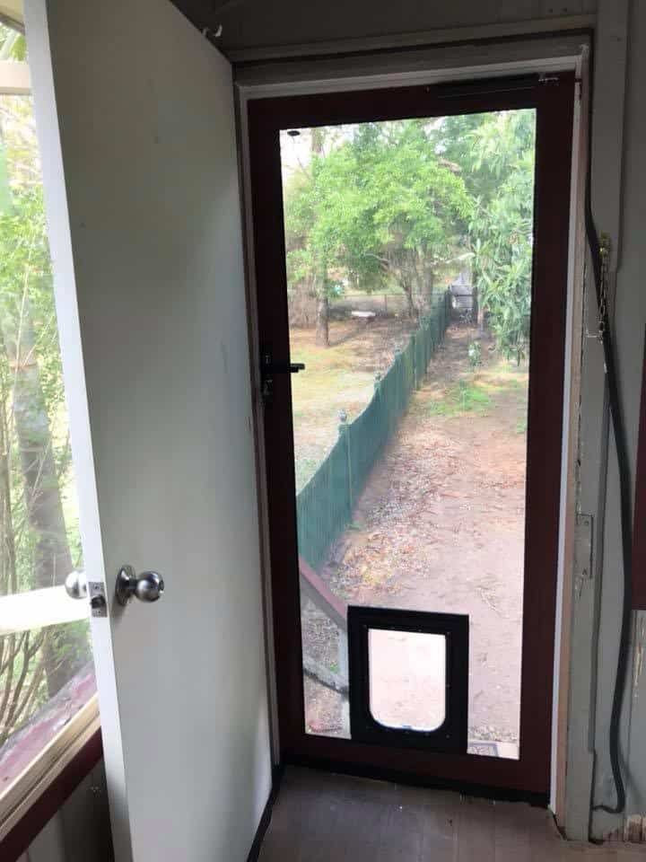An open white door reveals a brown screen door with a pet door installed at the bottom, looking out to a fenced yard.