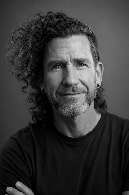 Black-and-white portrait of a man with curly hair and a beard, facing the camera with folded arms.