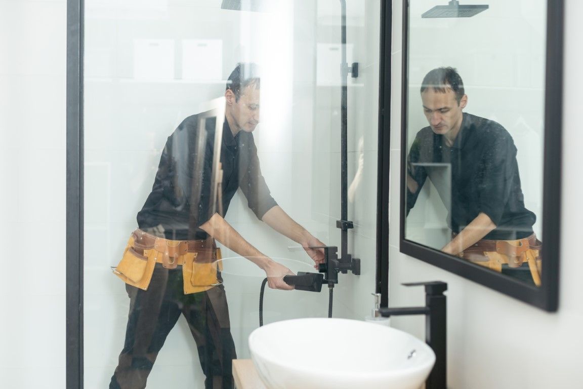 Plumber working on a shower fixture in a bathroom. He's holding a tool, wearing a tool belt, and is reflected in the mirror.