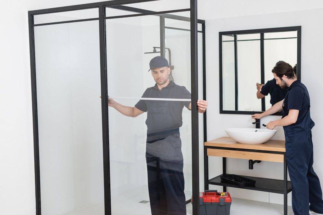 Two workers installing a shower enclosure in a bathroom. One measures, the other works on the sink.
