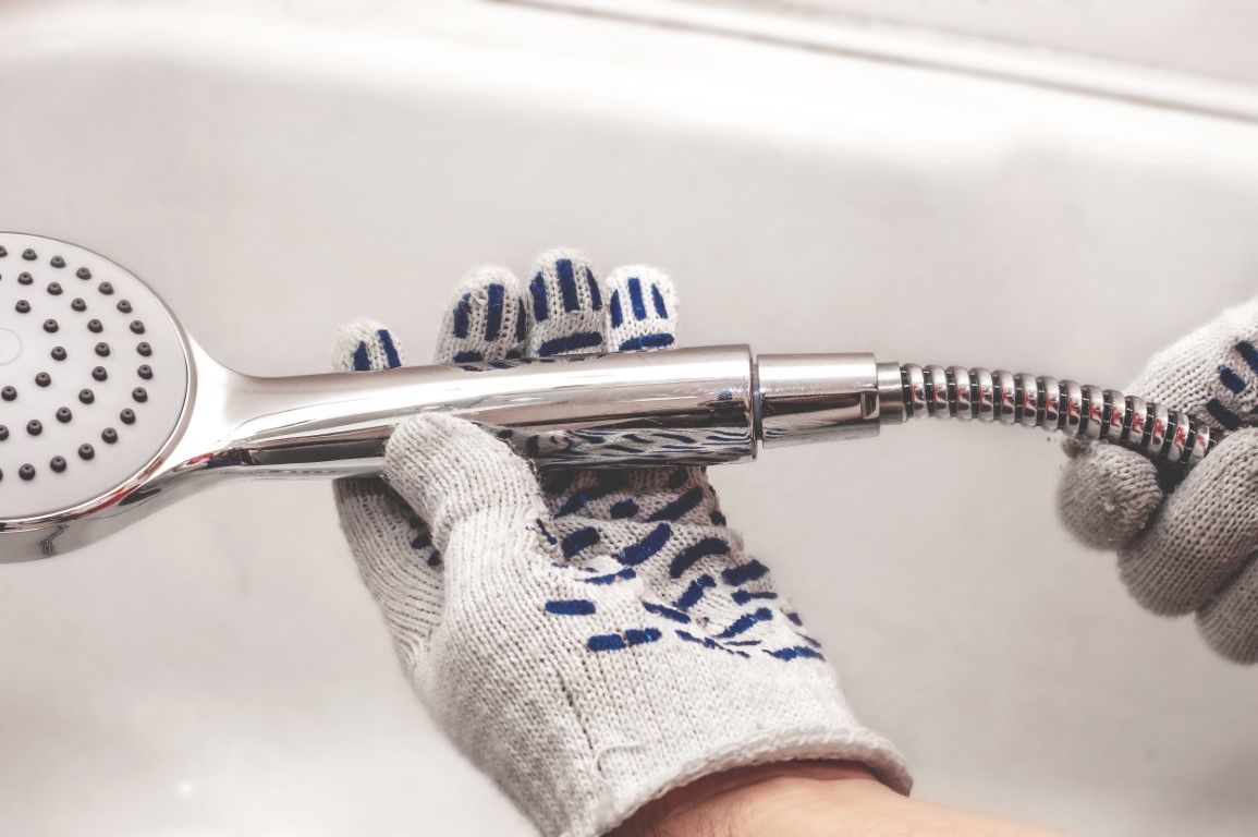 Hands wearing work gloves holding a silver showerhead and hose in a white tub.