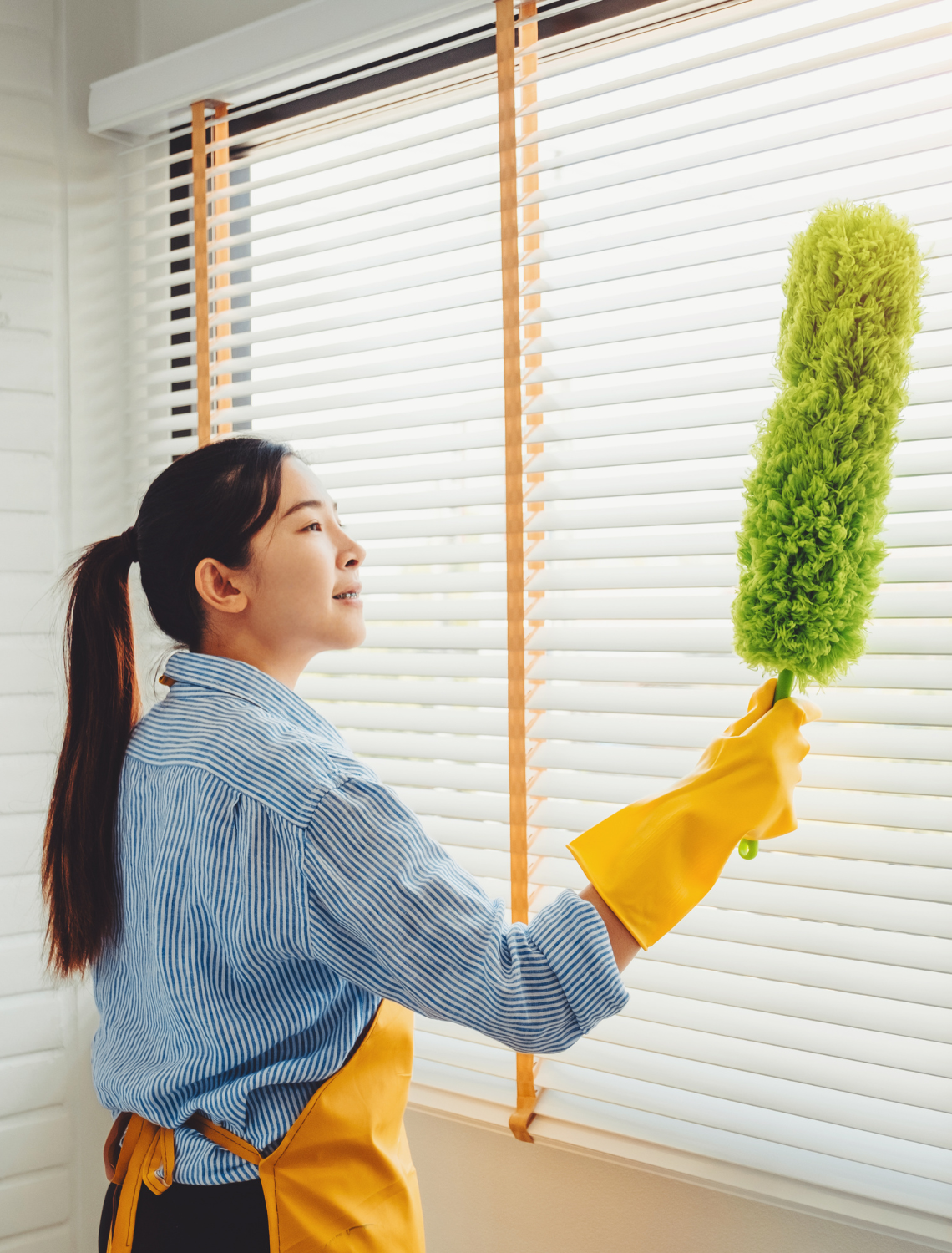 Woman in yellow gloves dusting blinds with a green duster beside a window