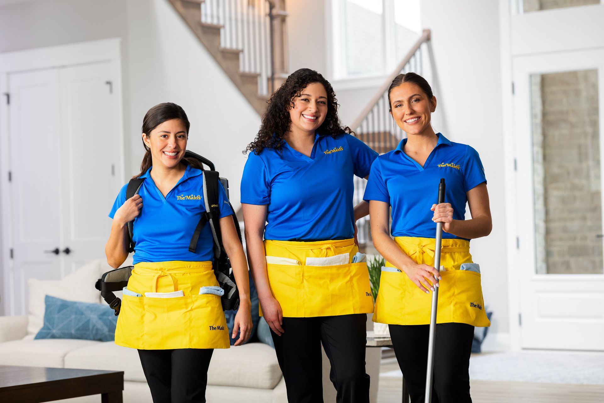 Three cleaners in blue shirts and yellow aprons holding mops in a bright living room