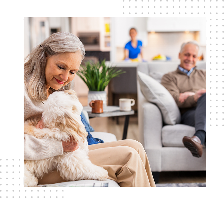 Woman cuddling a fluffy white dog in a bright living room, with a smiling man seated in the background