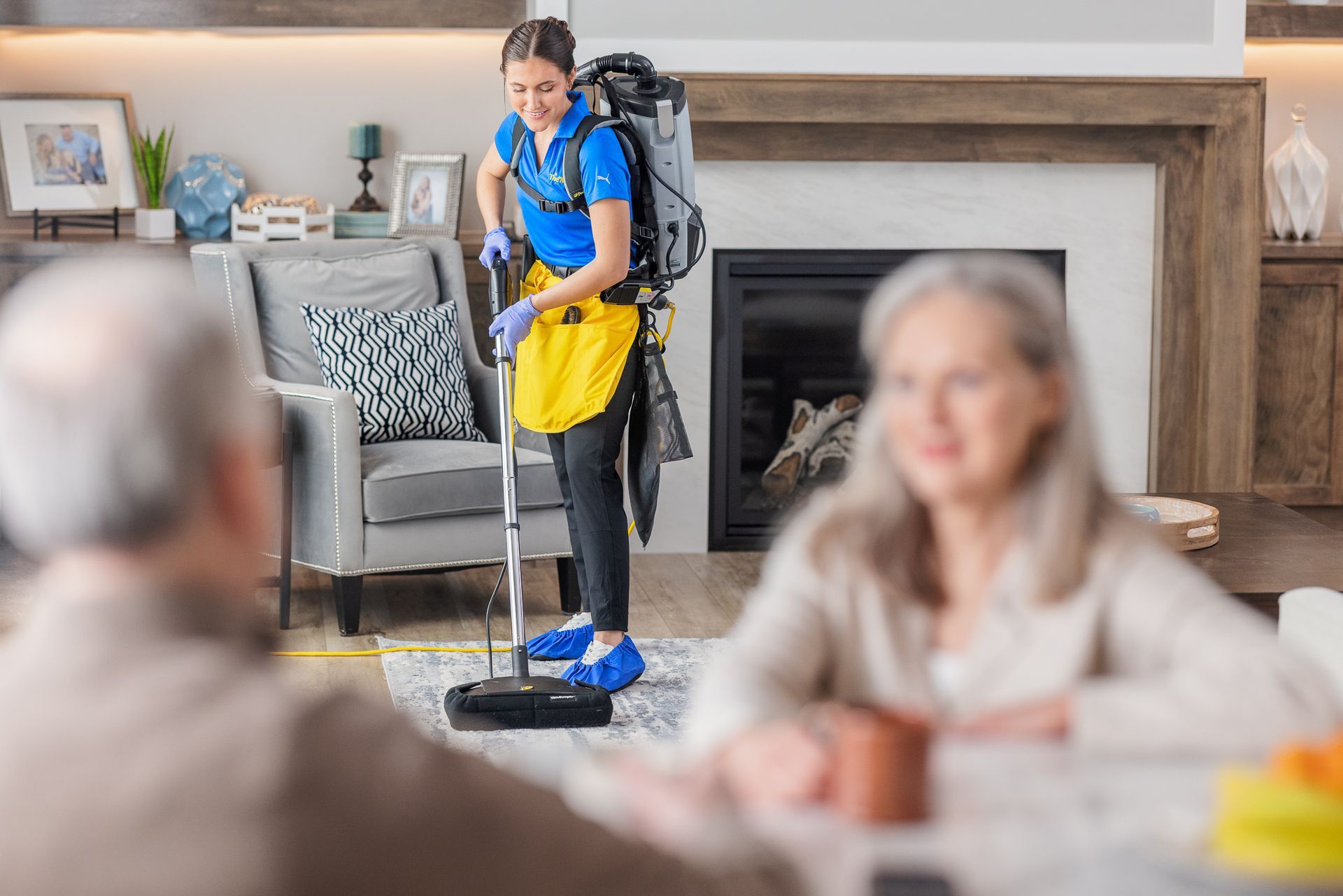 Housekeeper mopping a living room floor while two people sit blurred in the foreground