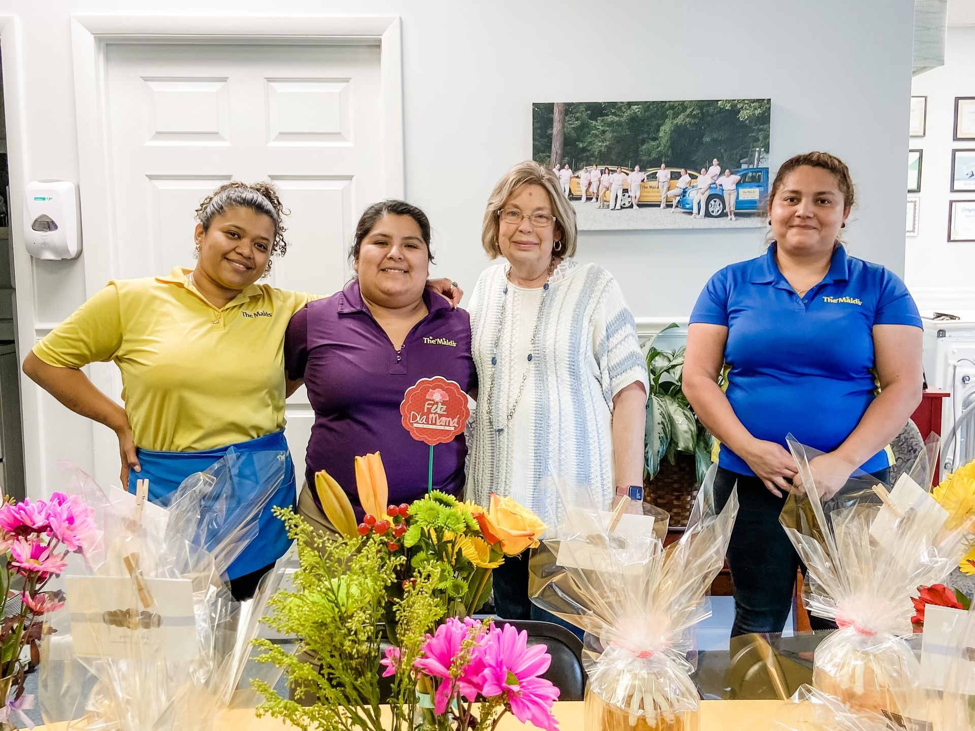 Five people stand behind a flower-covered table, posing indoors in front of a white door and wall art.