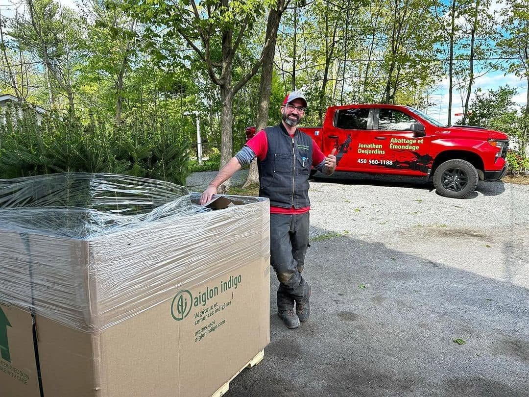Un homme debout près d’une grande palette filmée de plastique, à côté d’un pick-up rouge sur un chemin de gravier.
