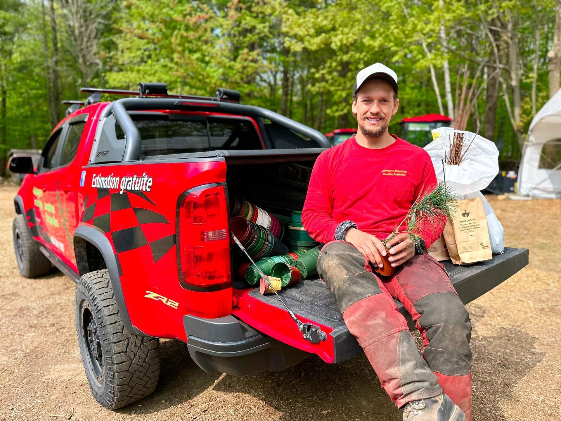 Un homme en chemise rouge assis sur le hayon d’un pick-up rouge, avec du matériel de chasse, dans une clairière en forêt