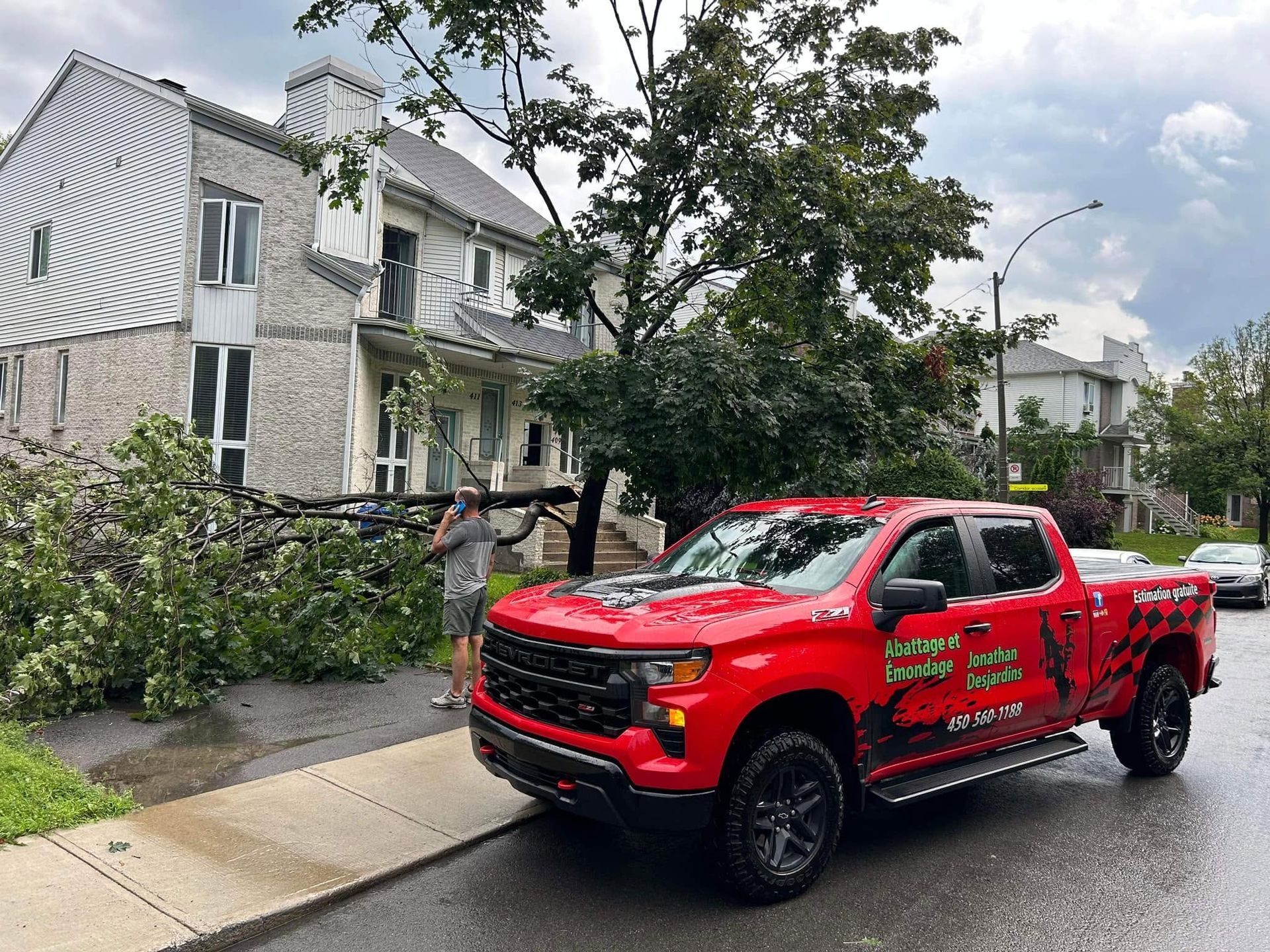 Camionnette rouge stationnée dans une rue mouillée, à côté d’une maison endommagée par la tempête, avec des branches d’arbre tombées à proximité