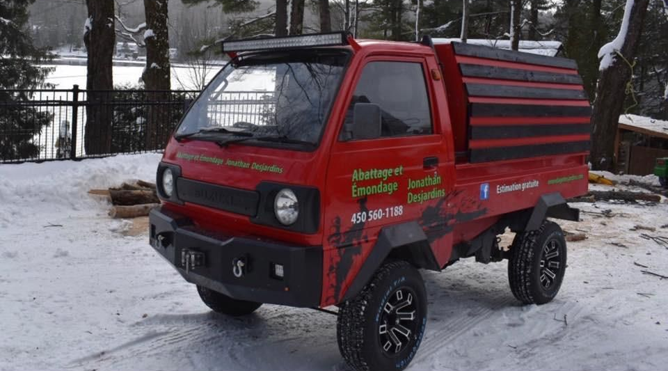 Petit camion utilitaire rouge avec galerie de toit noire, garé sur un sol enneigé près d’arbres et d’une clôture