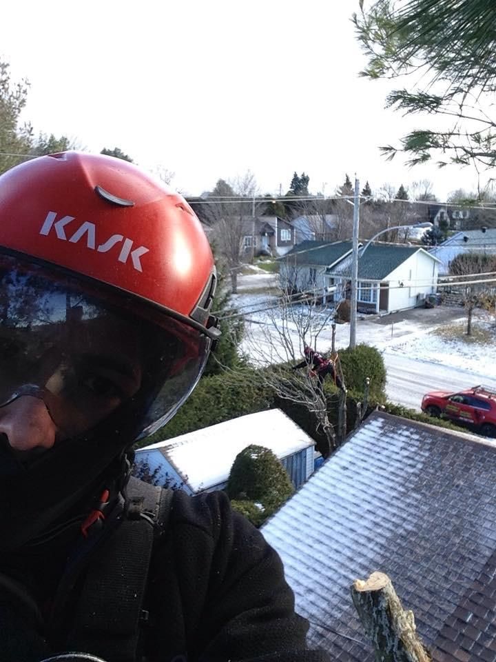 Une personne portant un casque orange prend un selfie sur un toit, avec un quartier résidentiel enneigé en arrière-plan.