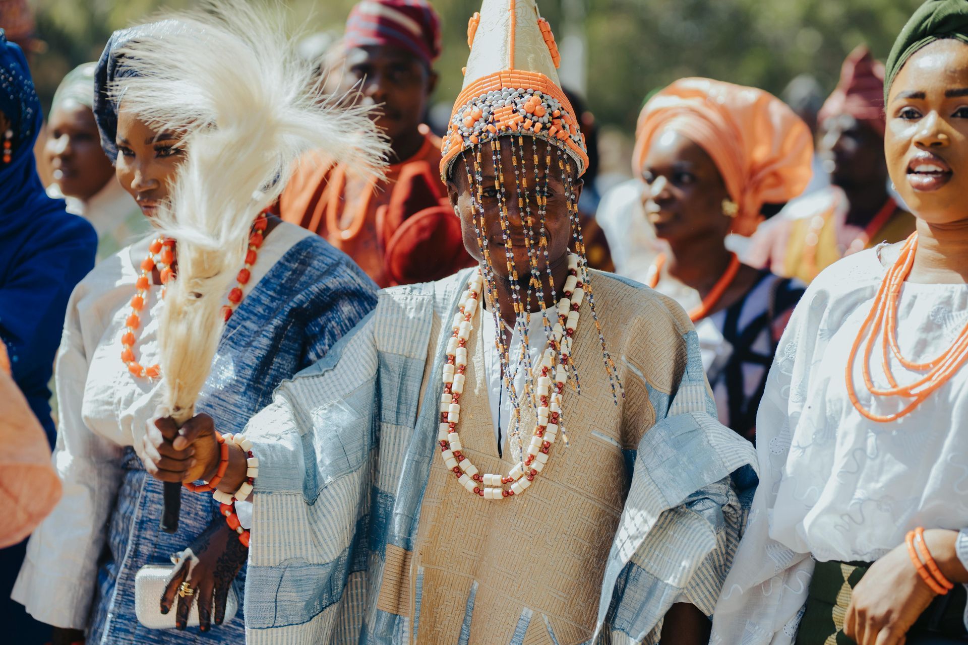 Group in traditional Nigerian attire, holding fan and beads, in outdoor setting.