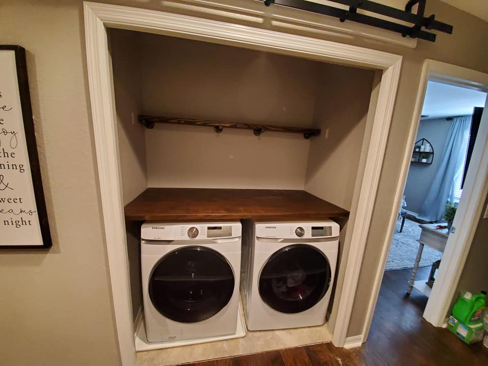 Laundry machines in a closet with a wooden countertop and shelf.