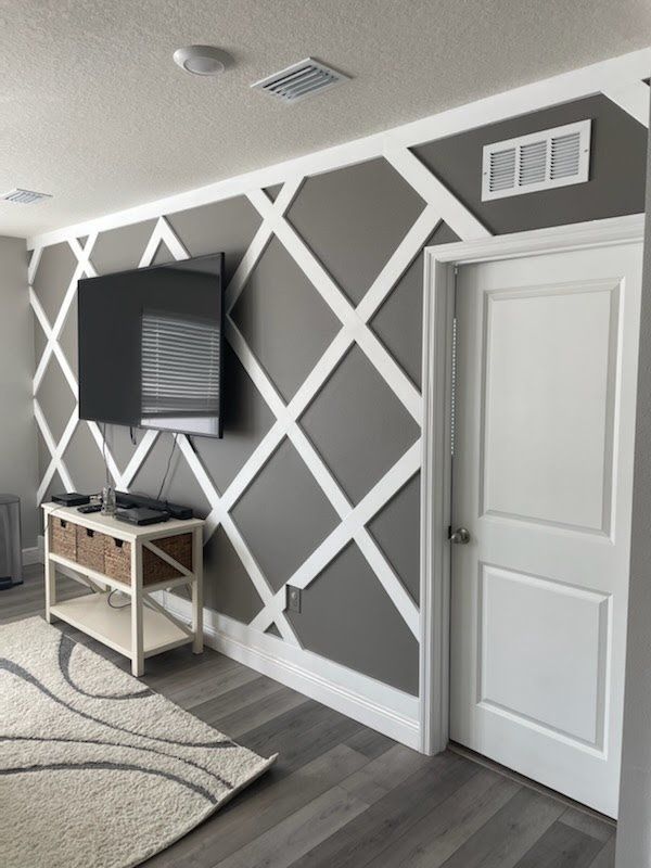 Gray accent wall with white diamond trim. TV and white door on the right. Entertainment stand with rug in foreground.