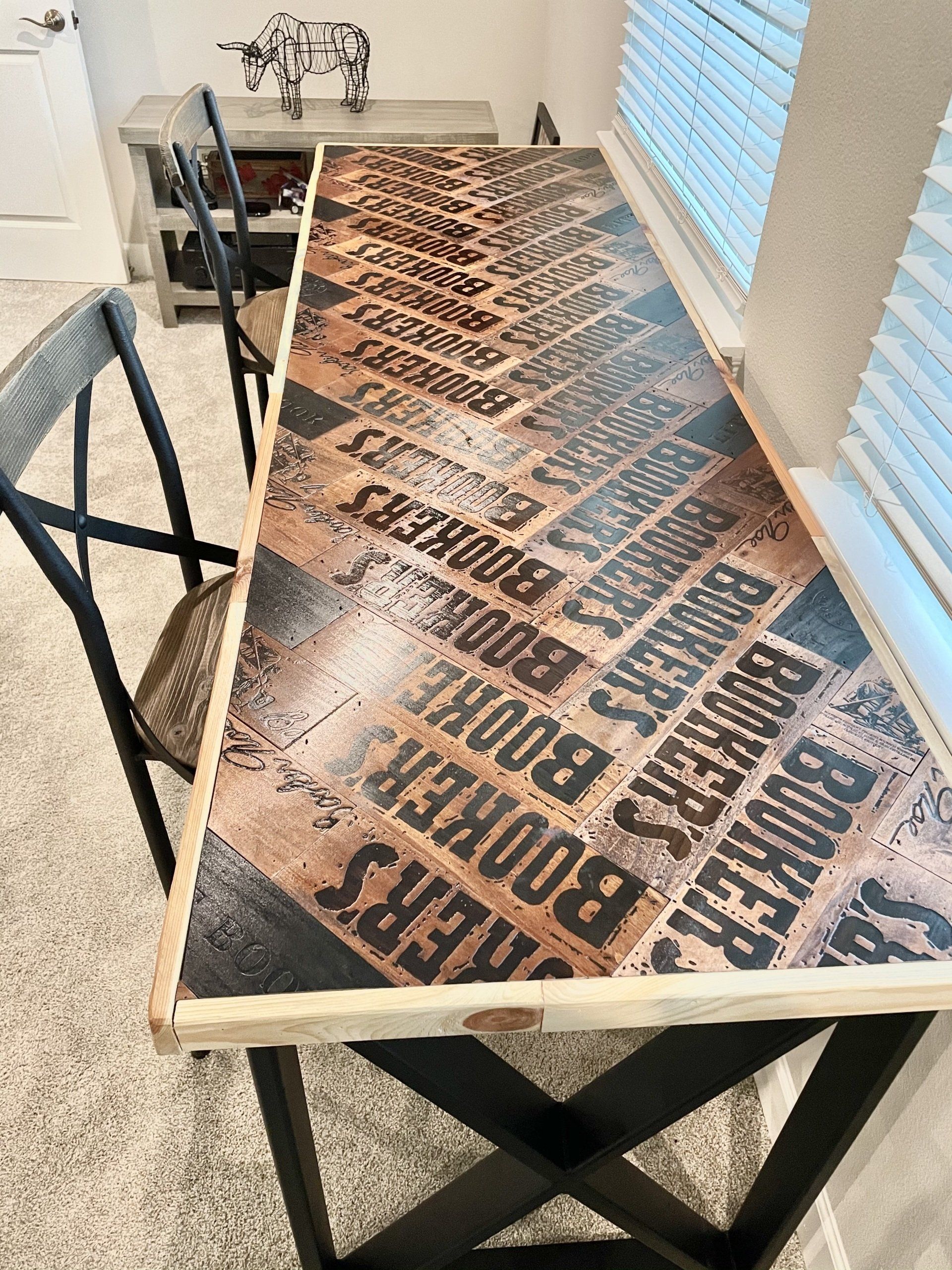 A narrow bar table with a whiskey barrel stencil top, black cross legs, and chairs, set in a room with a window.