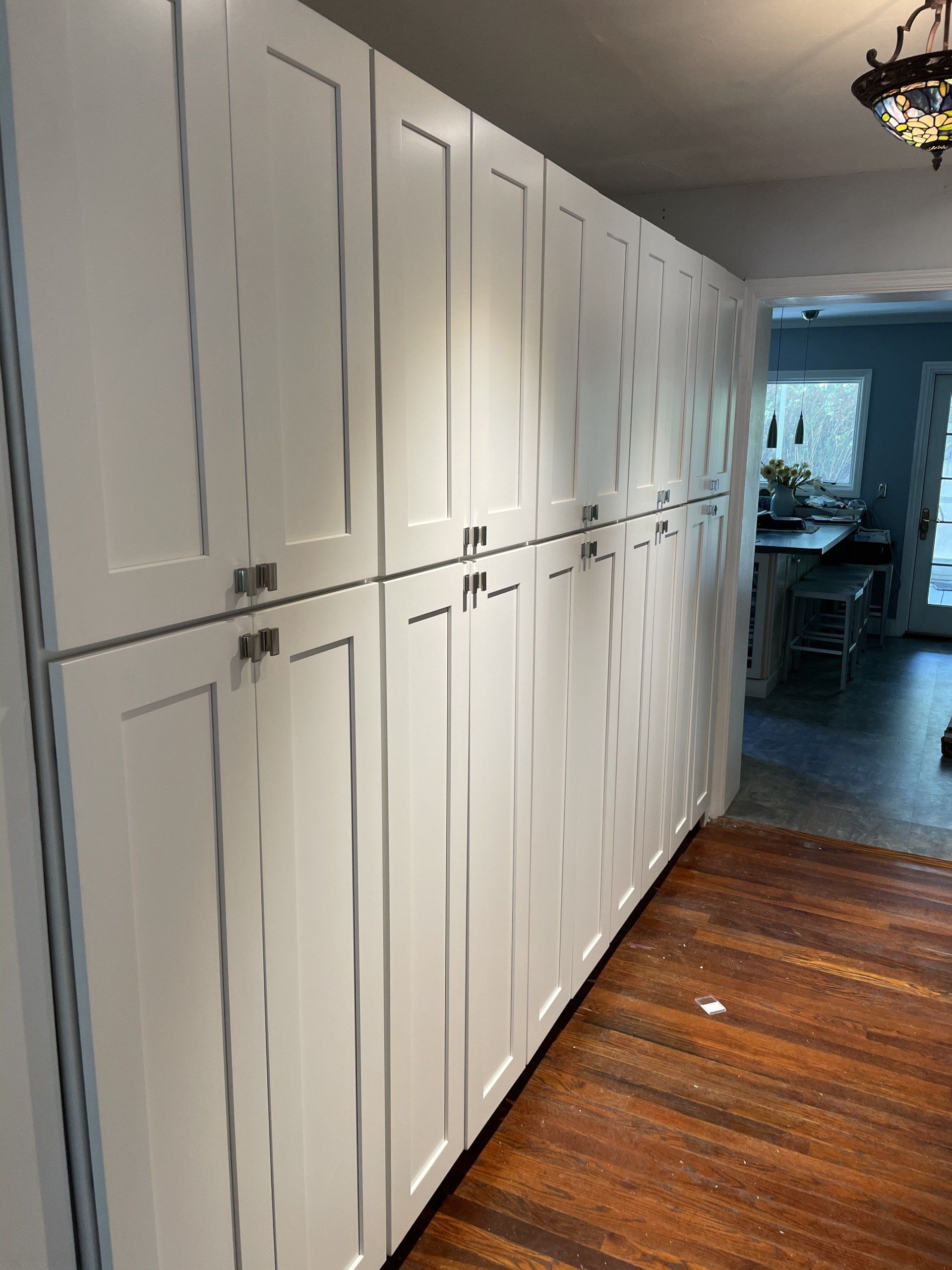 White pantry cabinets line a wall with silver handles, near a doorway to a kitchen with a wood floor.