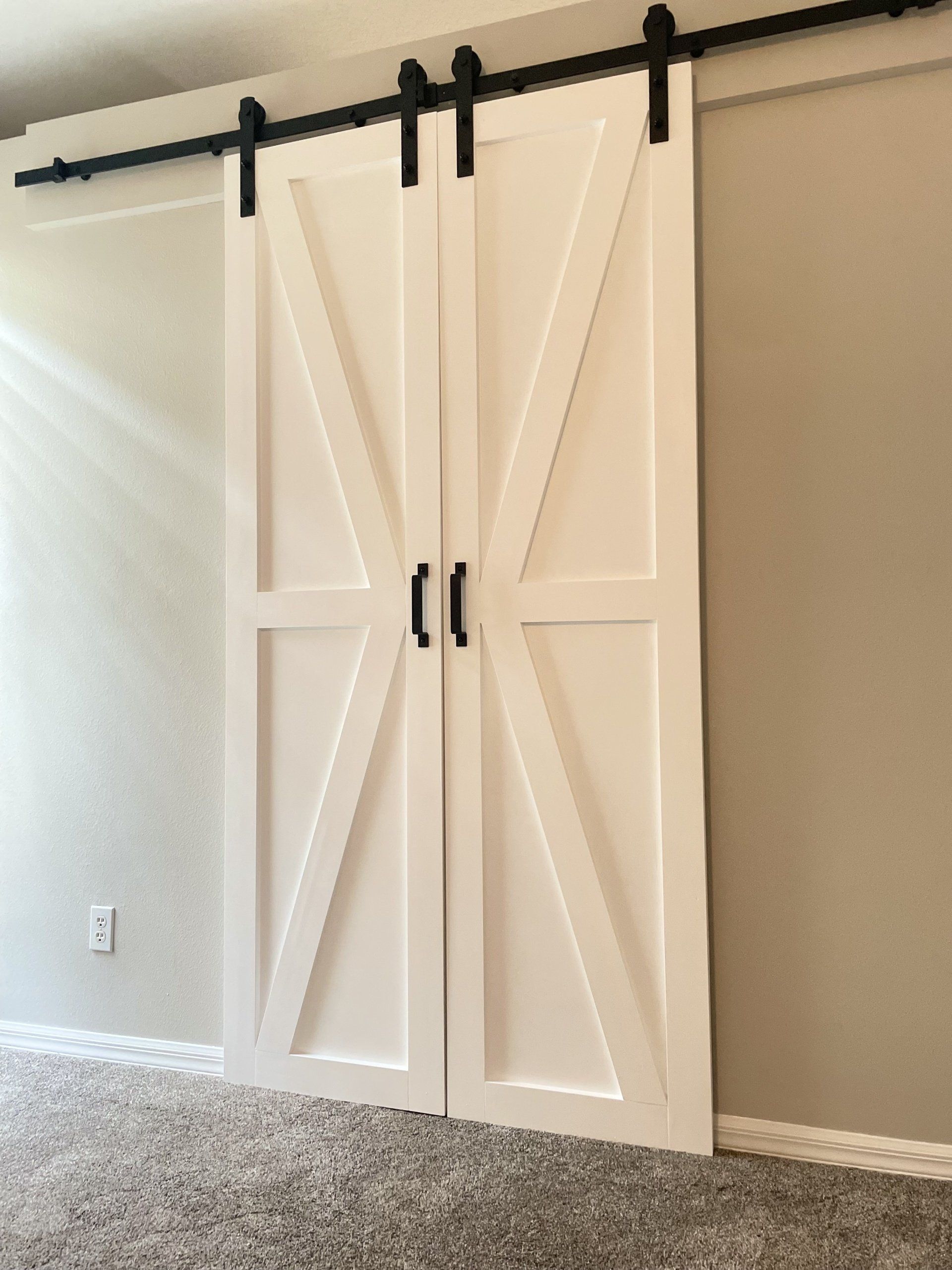 White barn doors with black hardware and handles on a beige wall, above a gray carpet.