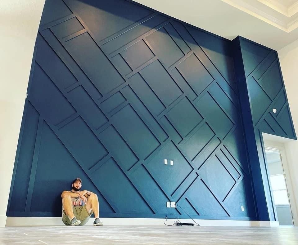 Man sits against a large navy blue accent wall with geometric molding. White trim and flooring.