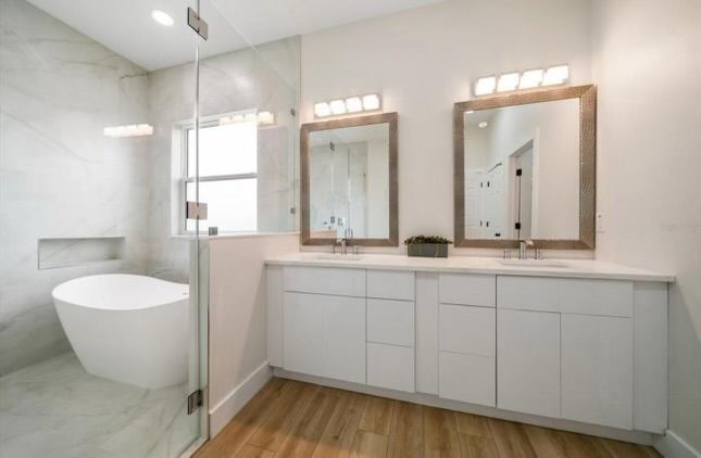 Modern bathroom with a white tub, vanity, and wood-look flooring. Mirrors and lights above the double sinks.