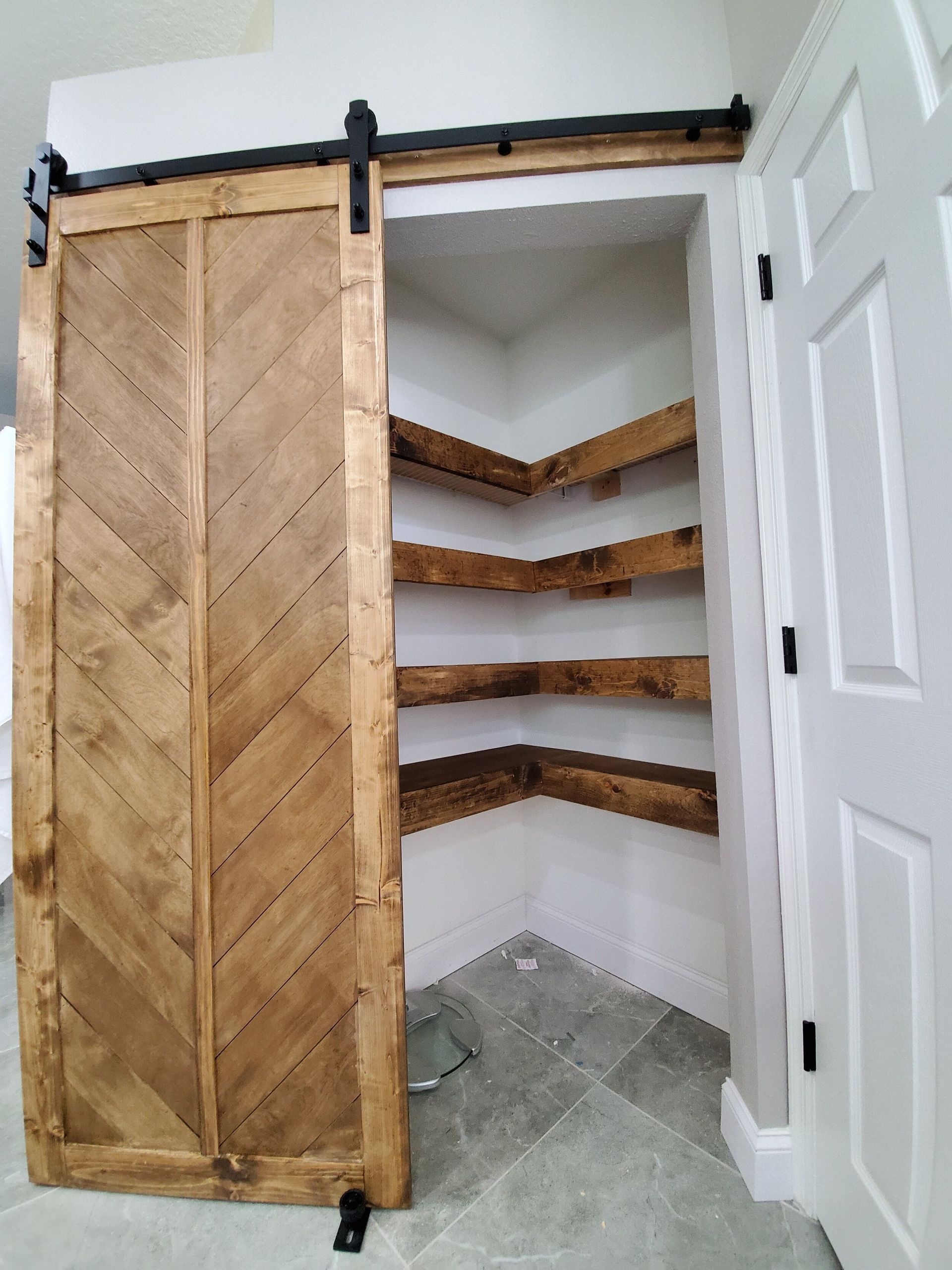 Barn door, chevron wood design, open to corner pantry with shelves. Light wood and white walls.