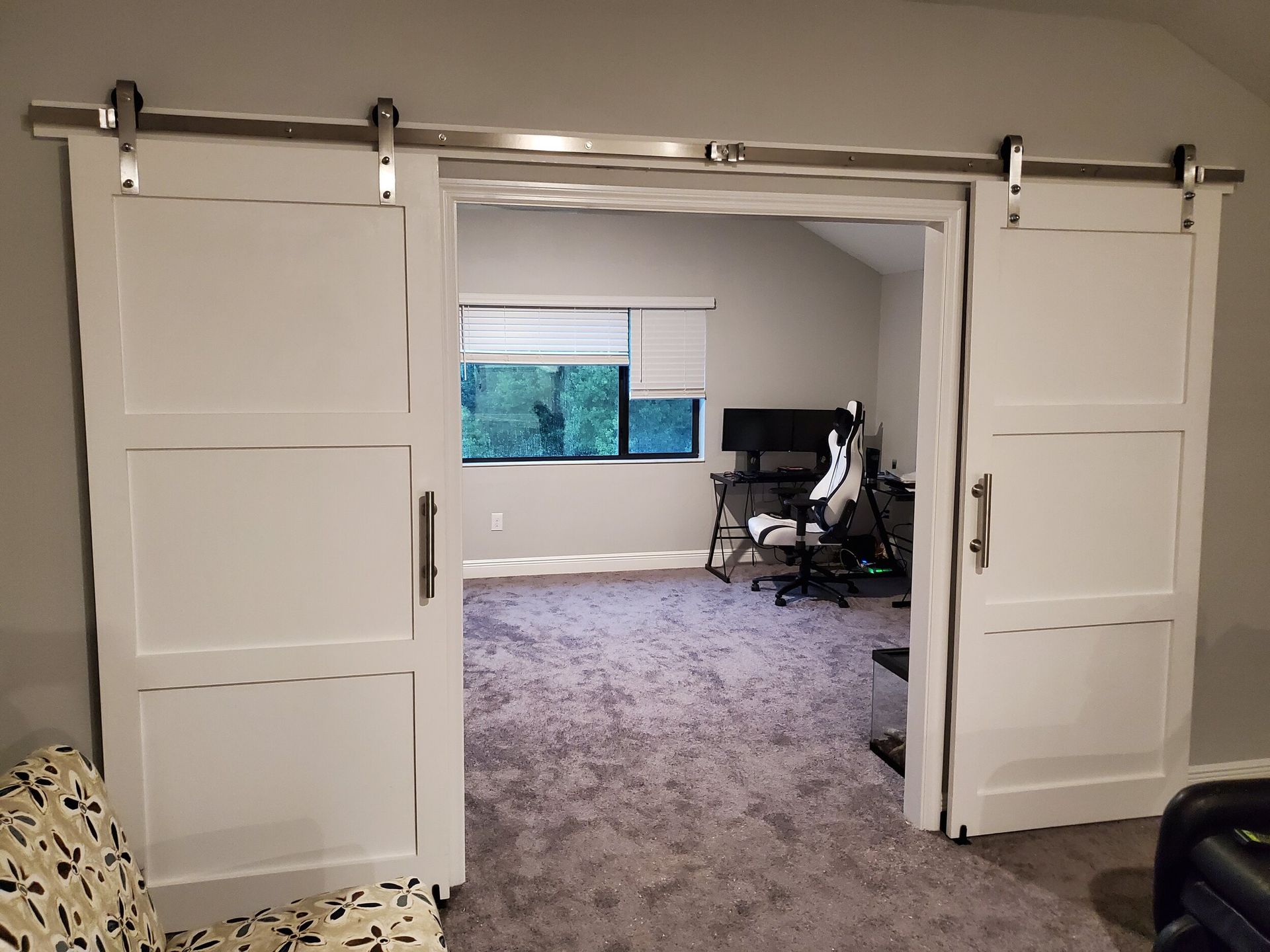 White sliding barn doors open to a room with a computer setup and window. Gray carpet.
