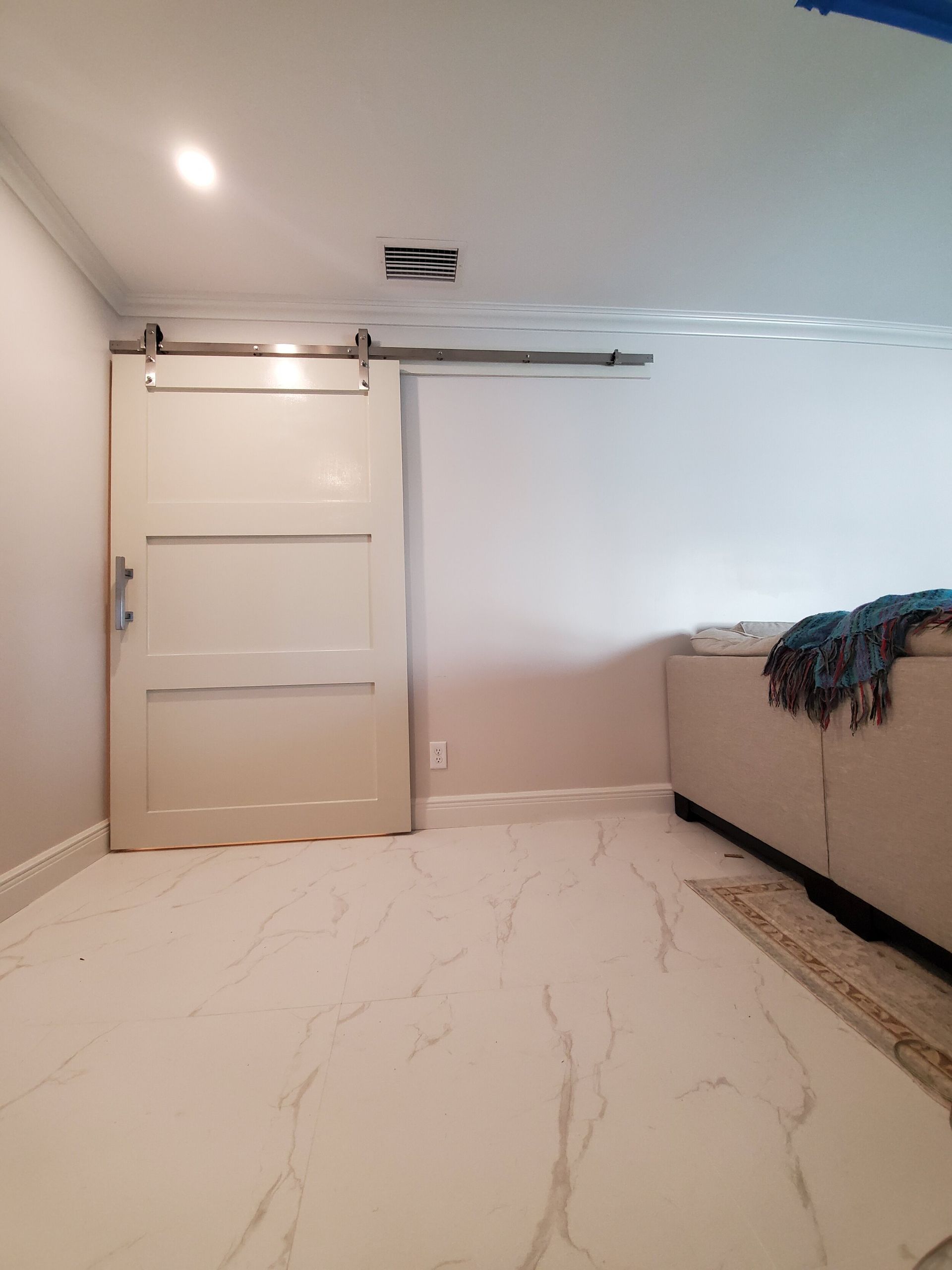 White barn door on a track, installed on a light-colored wall, in a room with light tile flooring and a sofa.