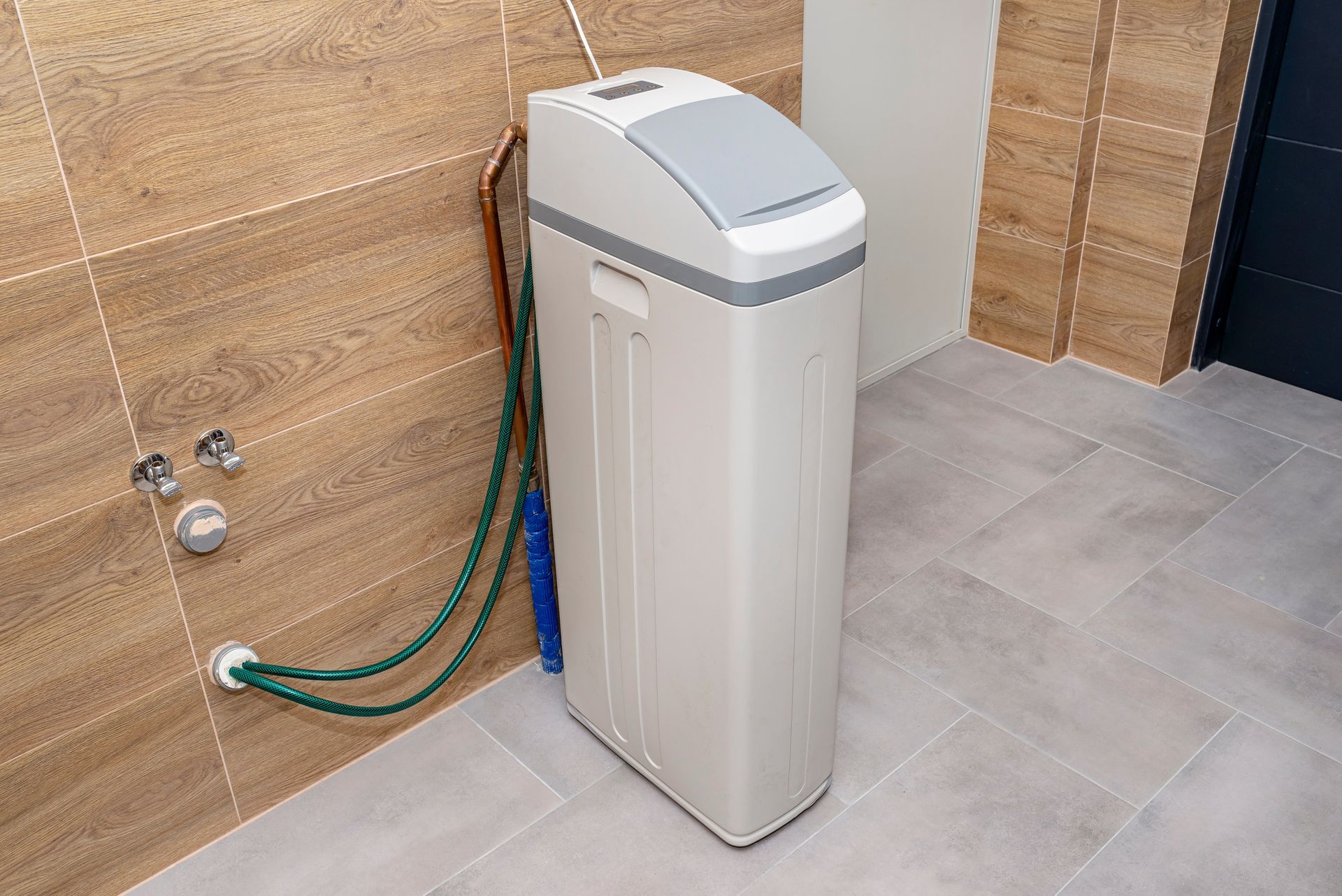 White residential water softener unit connected to copper piping and green hoses inside a tiled room