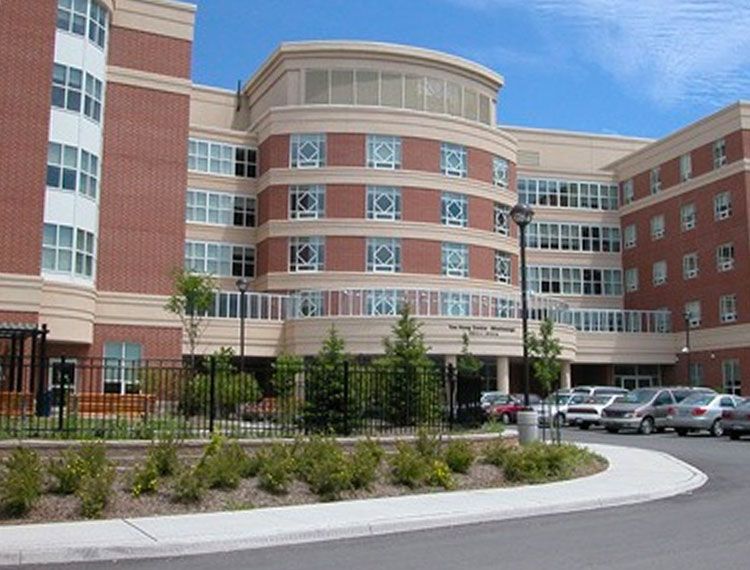 Brick and beige multi-story building with curved entrance, cars in a parking lot, blue sky.