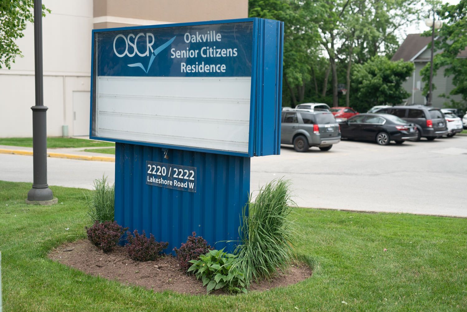Sign for Oakville Senior Citizens Residence in front of a building and parked cars; blue sign with white lettering.