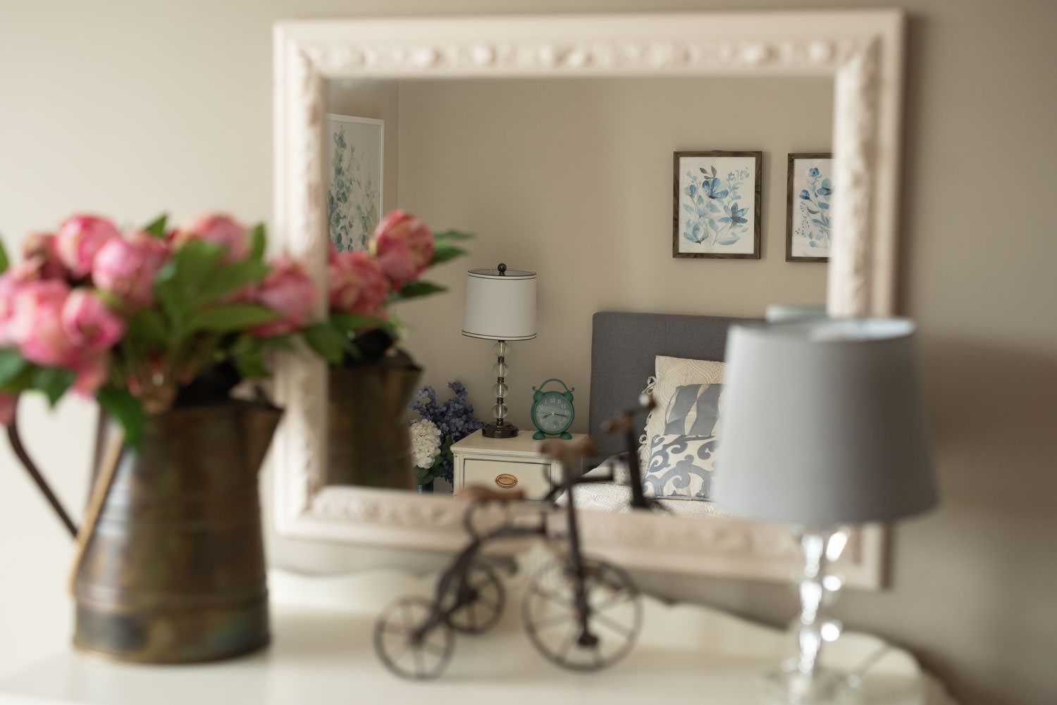 Rustic vase with pink flowers on a white dresser, reflected in a framed mirror, showing a bedroom with artwork.