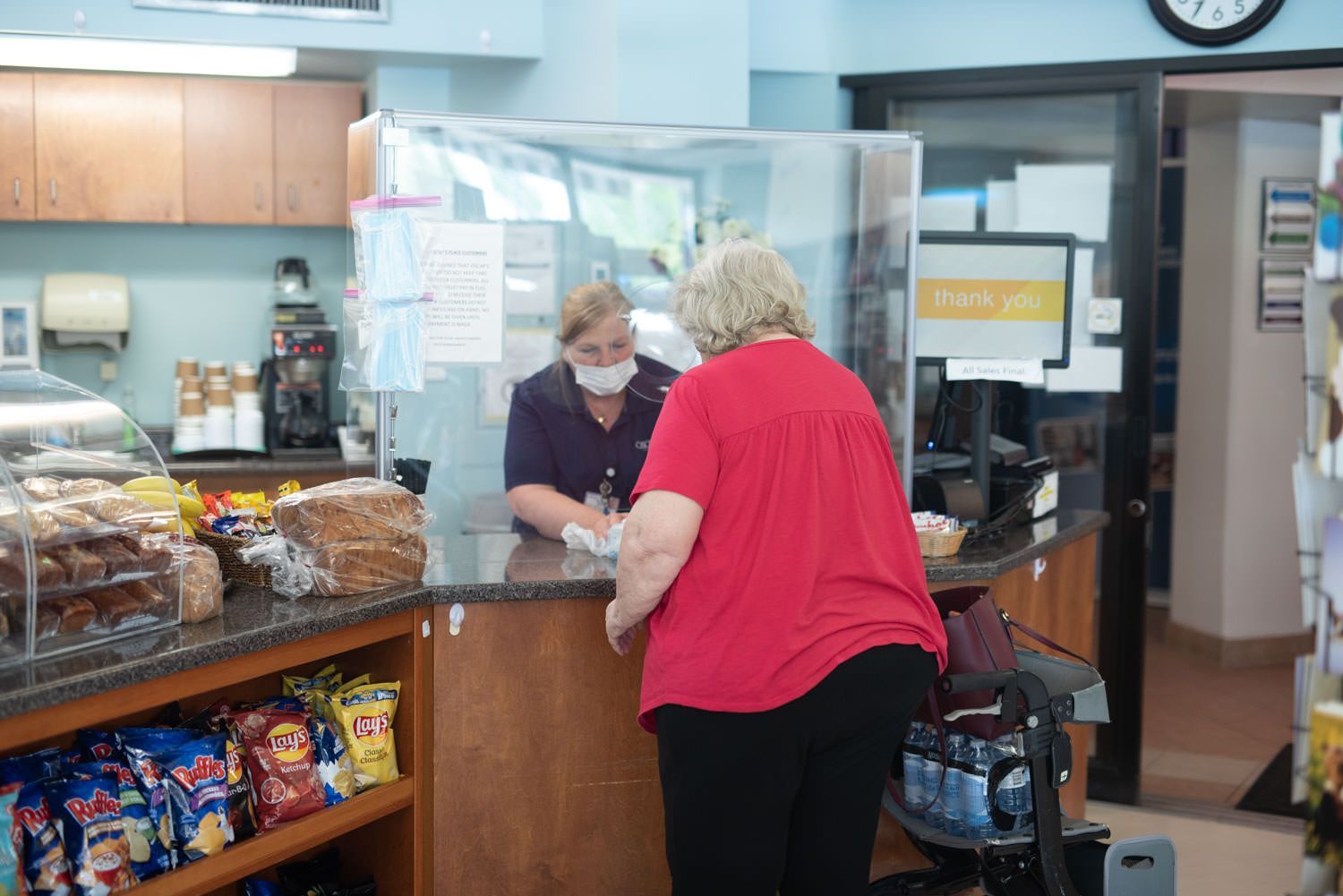 A masked person serves a customer behind a plexiglass barrier at a cafe counter with snacks and drinks.