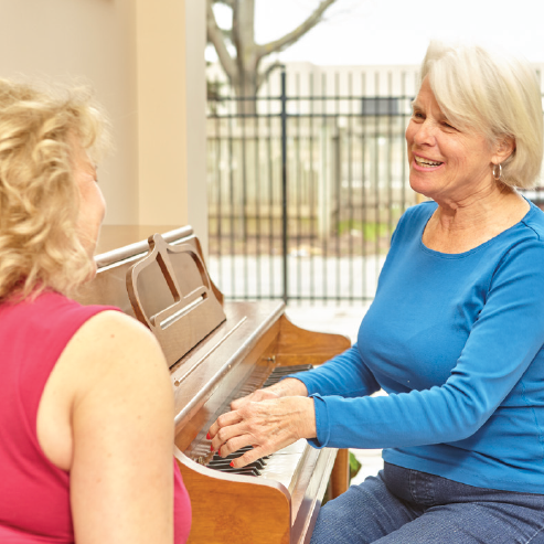 Woman plays piano while another woman listens. Light-colored hair, blue top, piano indoors.