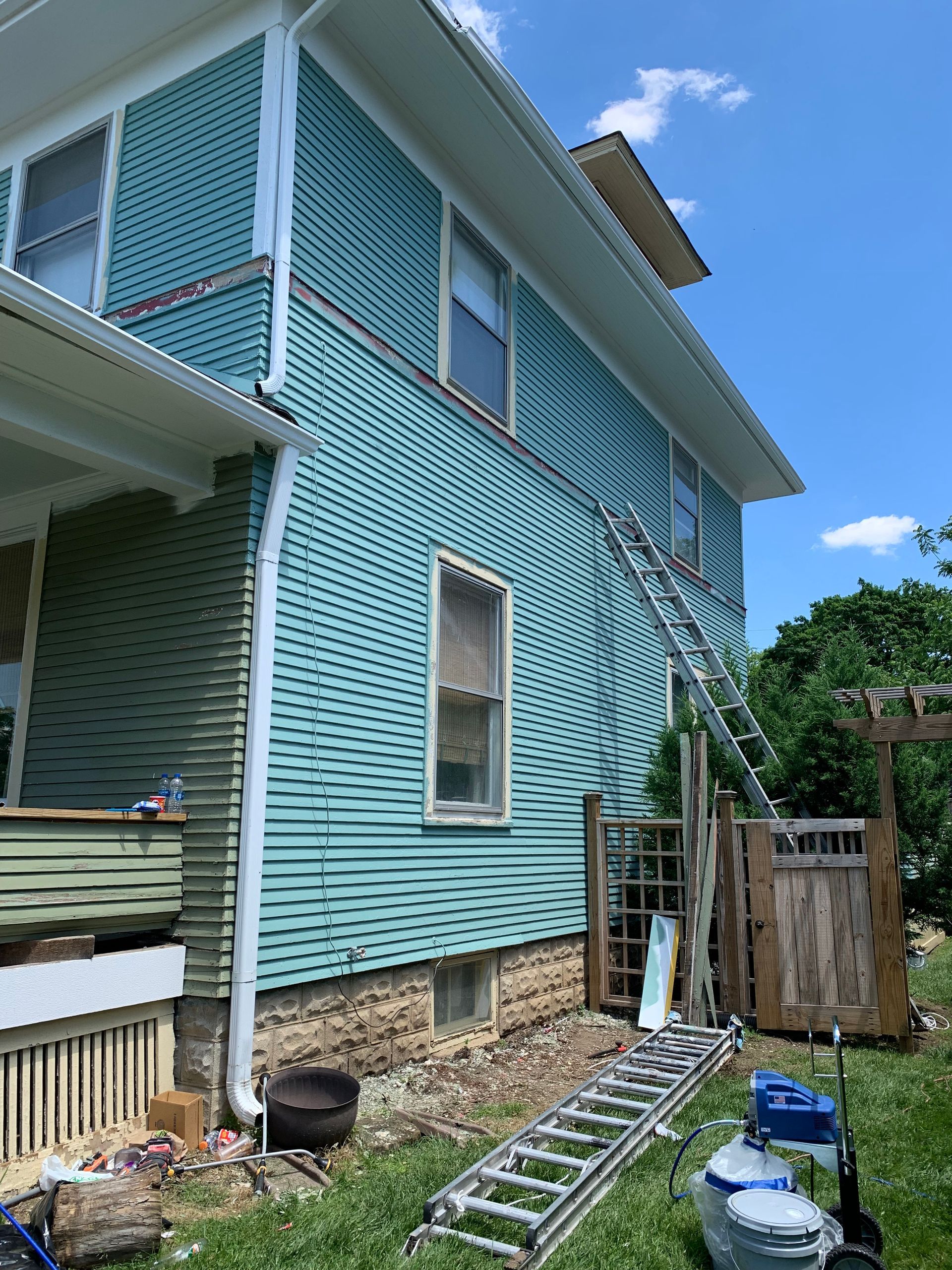 Light blue house exterior with white lattice siding, ladder, and yard tools along the foundation