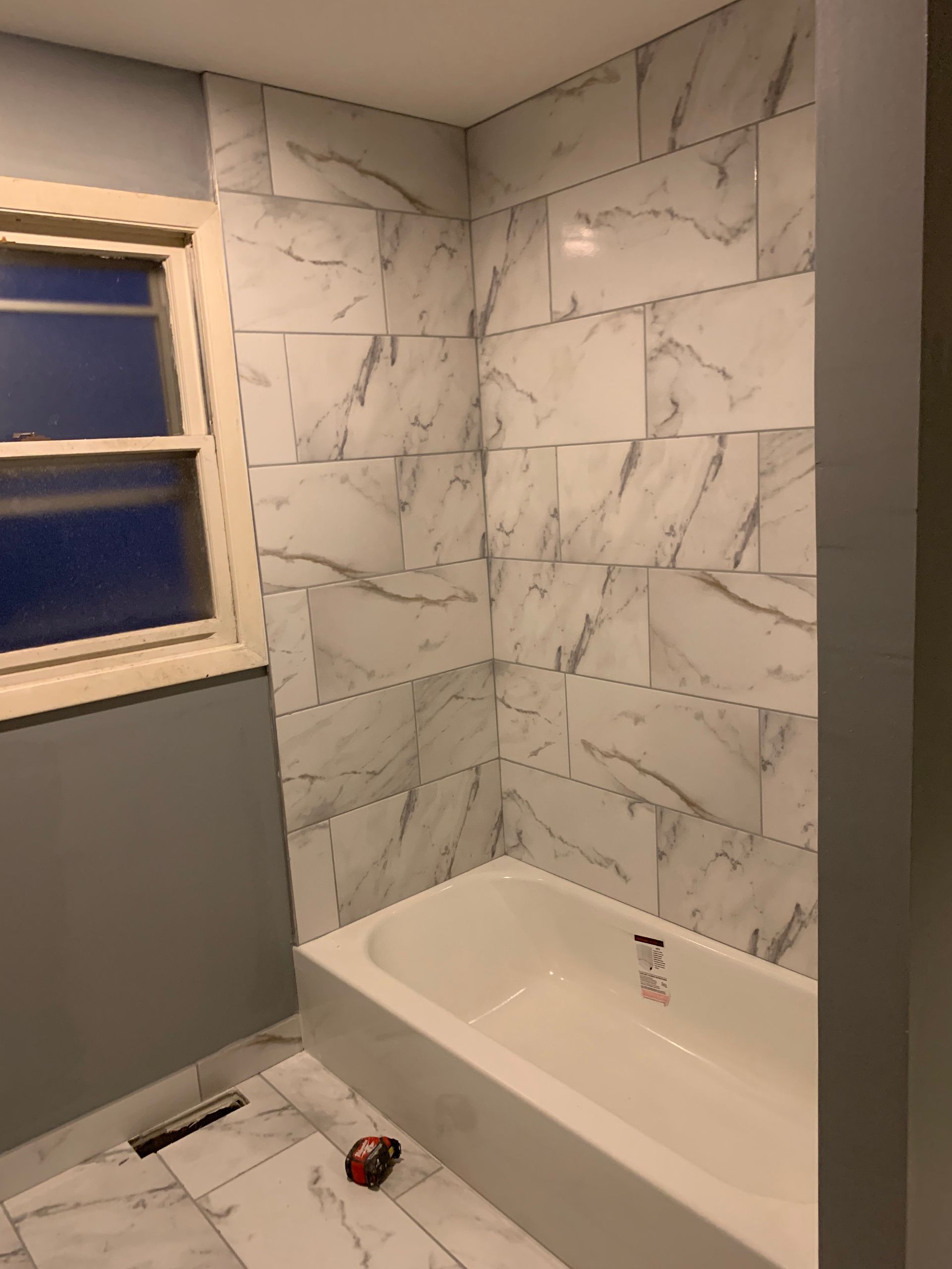 Bathroom with white marble-look shower walls, gray walls, and a white tub beside a window
