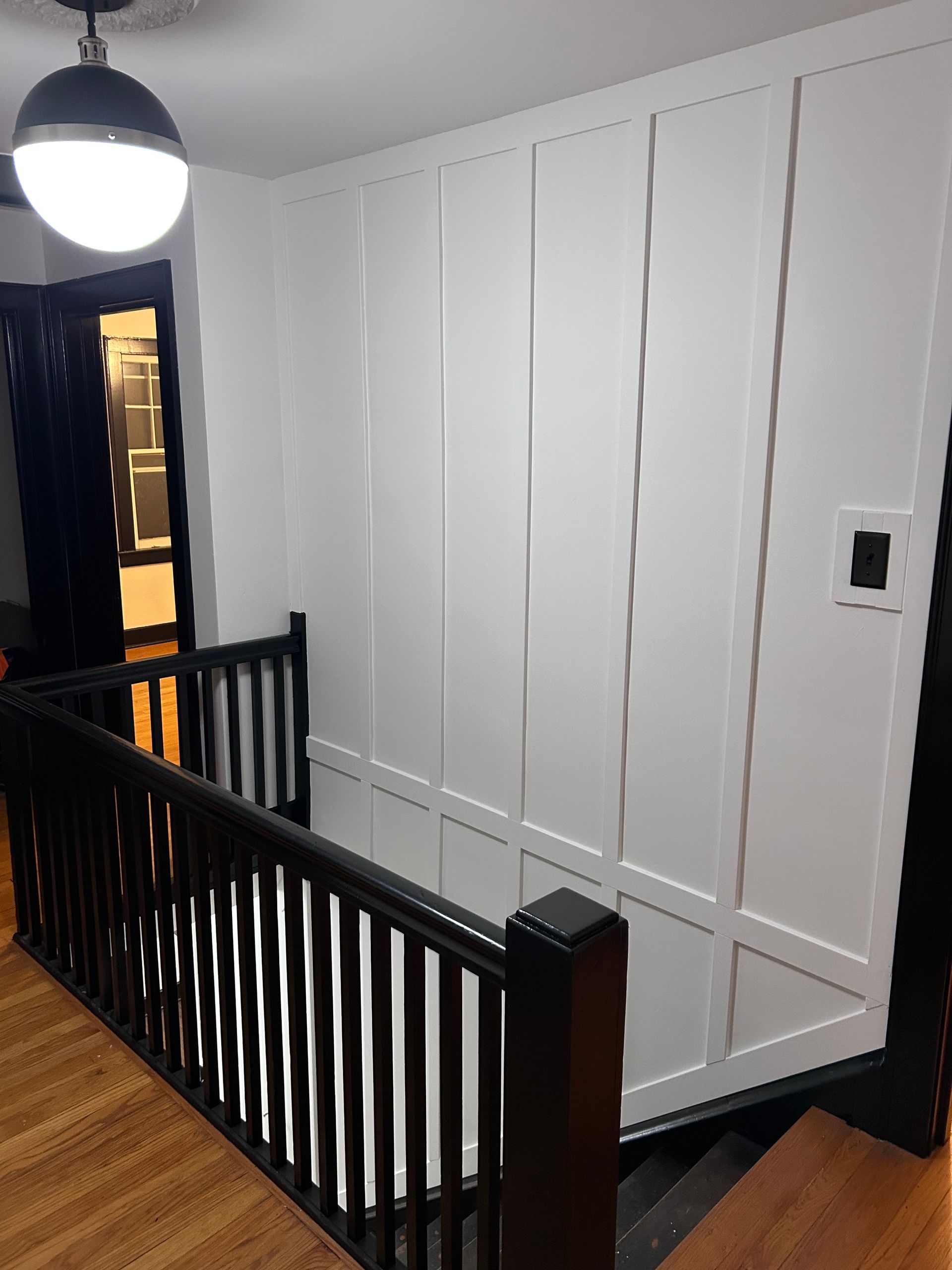 White paneled wall and black railing in a stairwell landing with wood floor and hanging globe light.