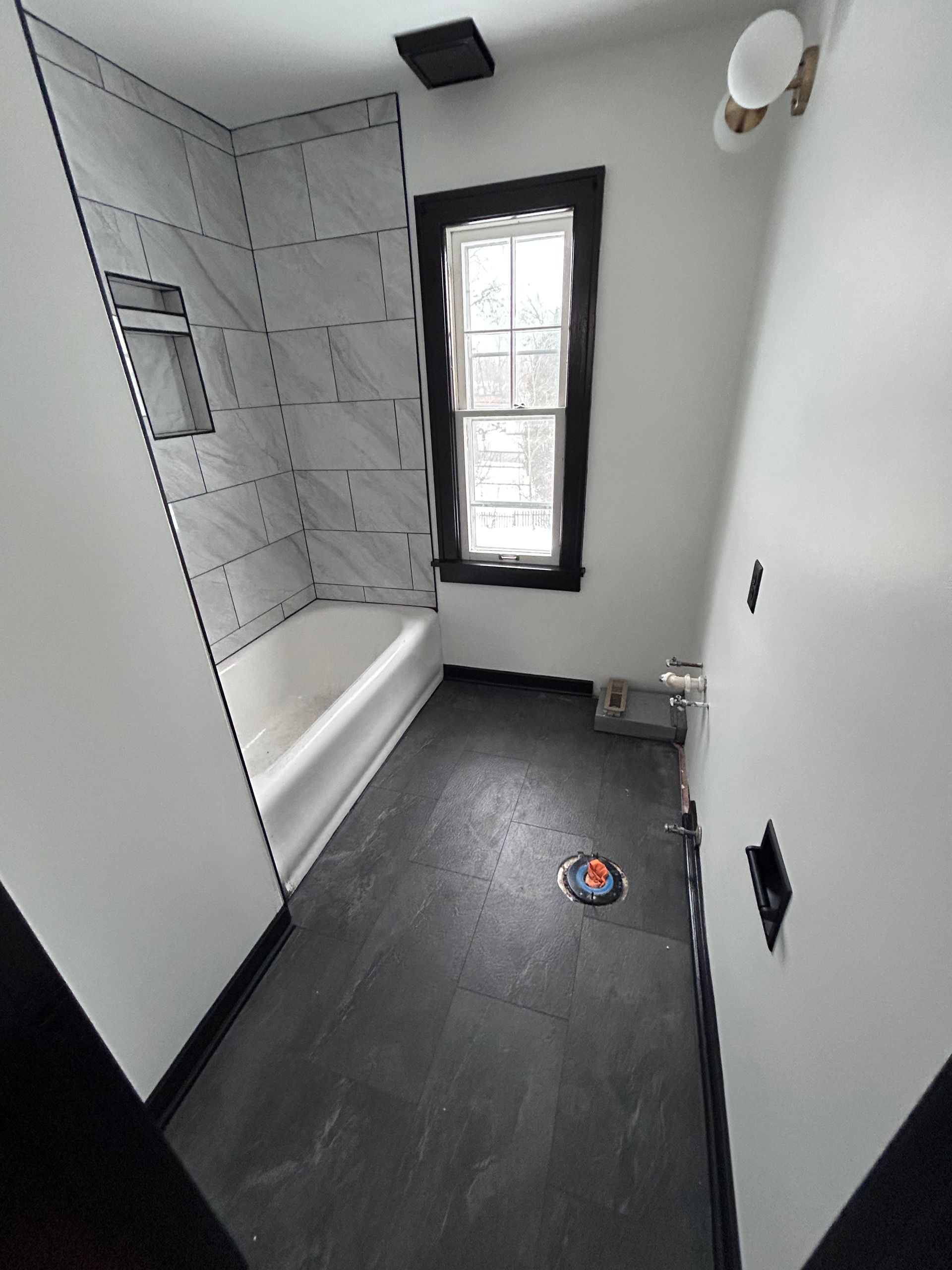 Narrow bathroom with bathtub, window, and dark floor under renovation.