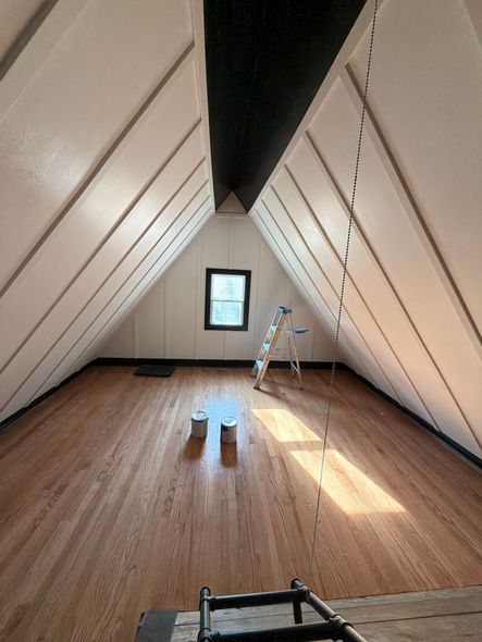 Empty attic room with sloped white walls, wood floor, a small window, and a ladder near the back wall