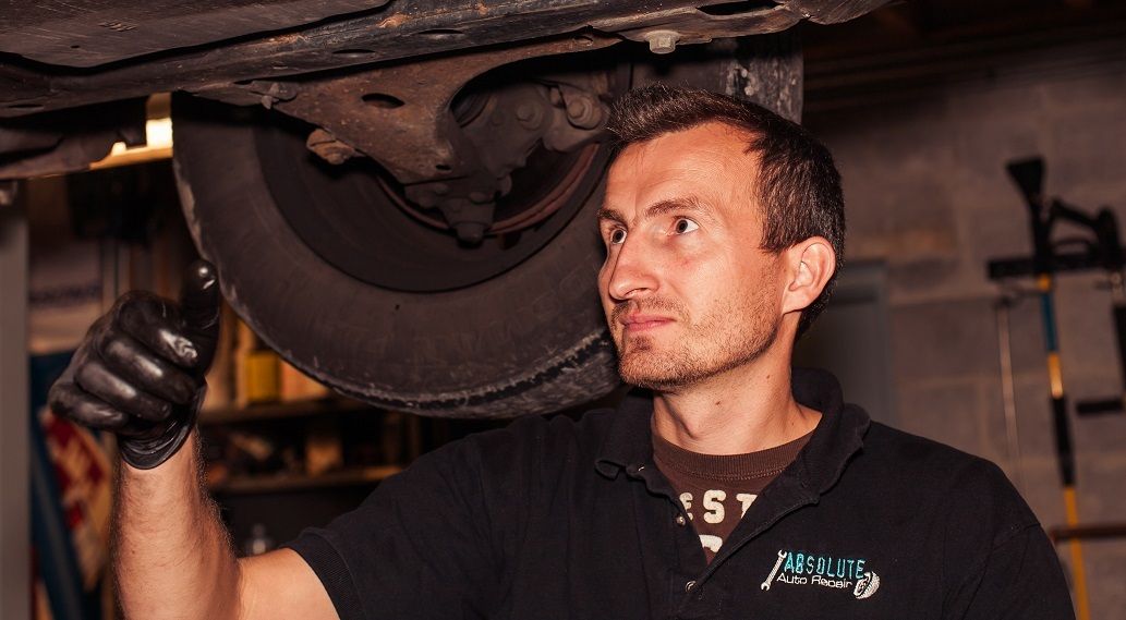Mechanic inspecting a car's underside with a flashlight, wearing gloves and a black shirt in a garage setting. | Absolute Auto Repairs & Sales