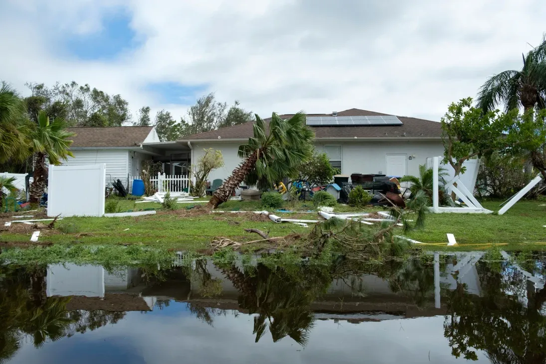 A flooded residential yard with downed palm trees and damaged white fencing in front of a house after a storm.