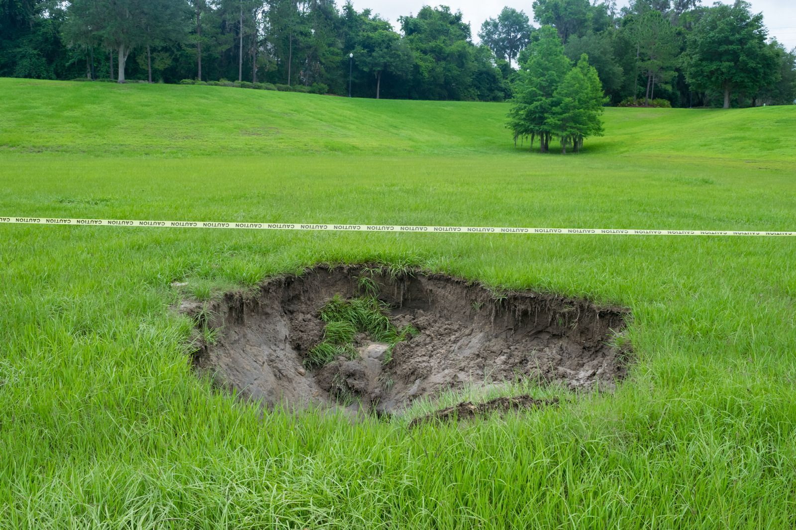Large sinkhole in a grassy field with exposed dirt and trees in the background