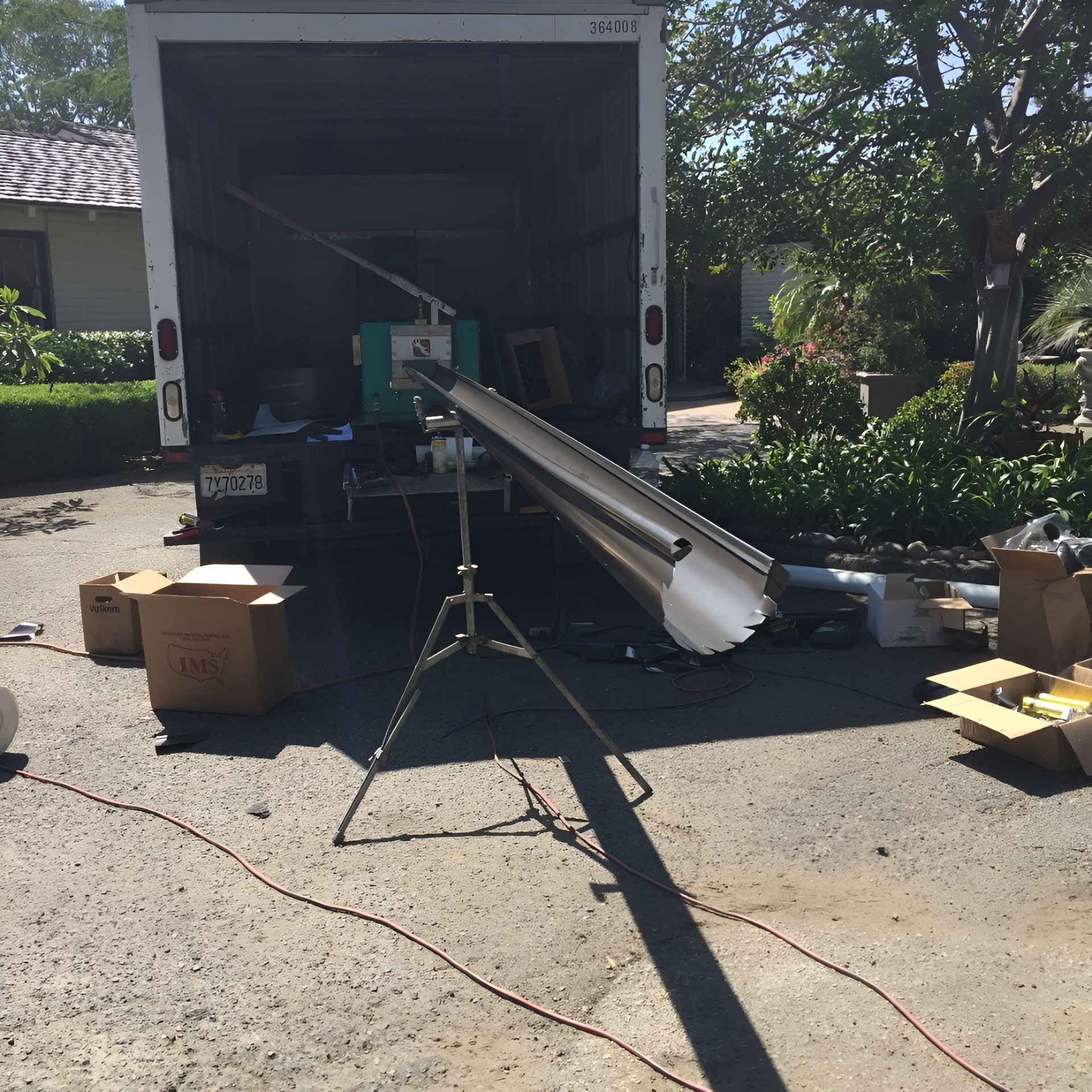 Open truck with a long metal beam extending out, boxes on the driveway, and a tripod in front.