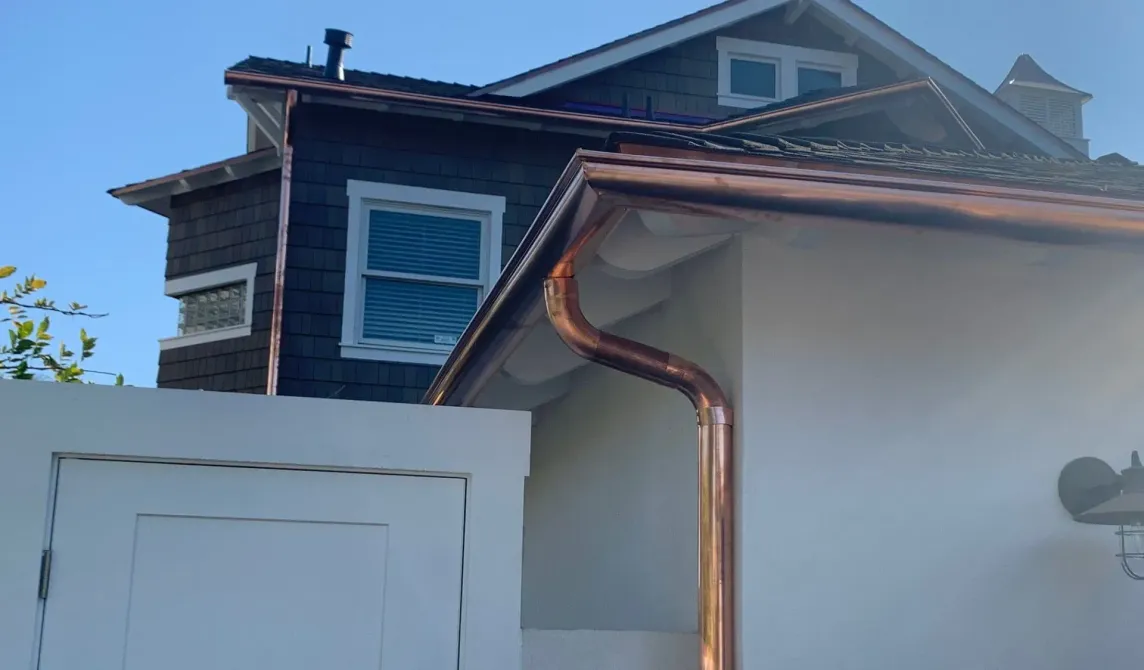 Blue house roofline with copper downspout above a white fence on a sunny day