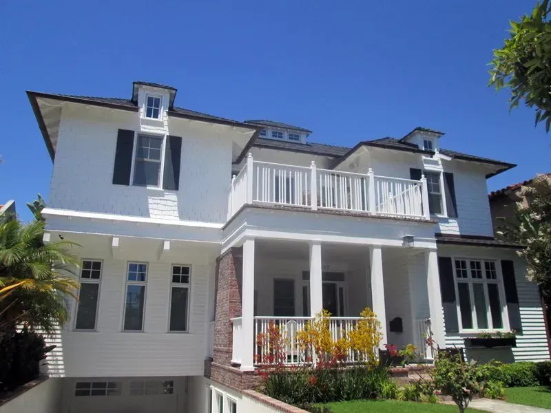 Large white two-story house with a balcony, columns, and a front garden under a blue sky