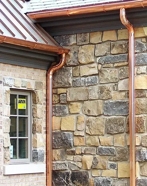 Stone exterior wall with copper downspouts and a small window under a roof overhang