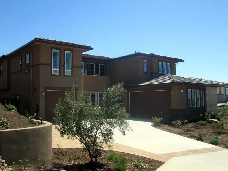 Modern two-story brown house with a driveway, garage, and small tree under a clear blue sky