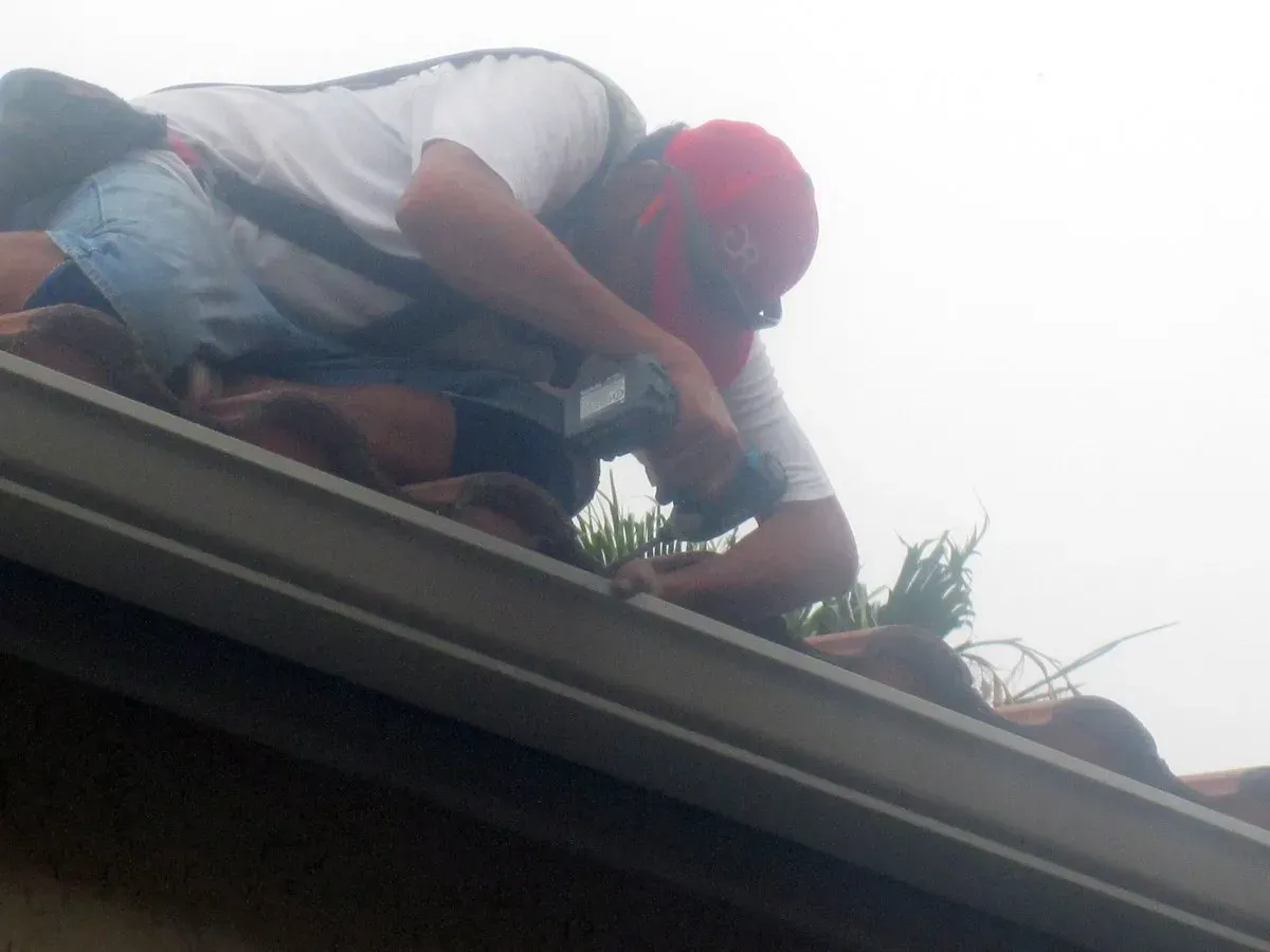 Worker crouching on a roof, handling tools near the edge under a bright sky.