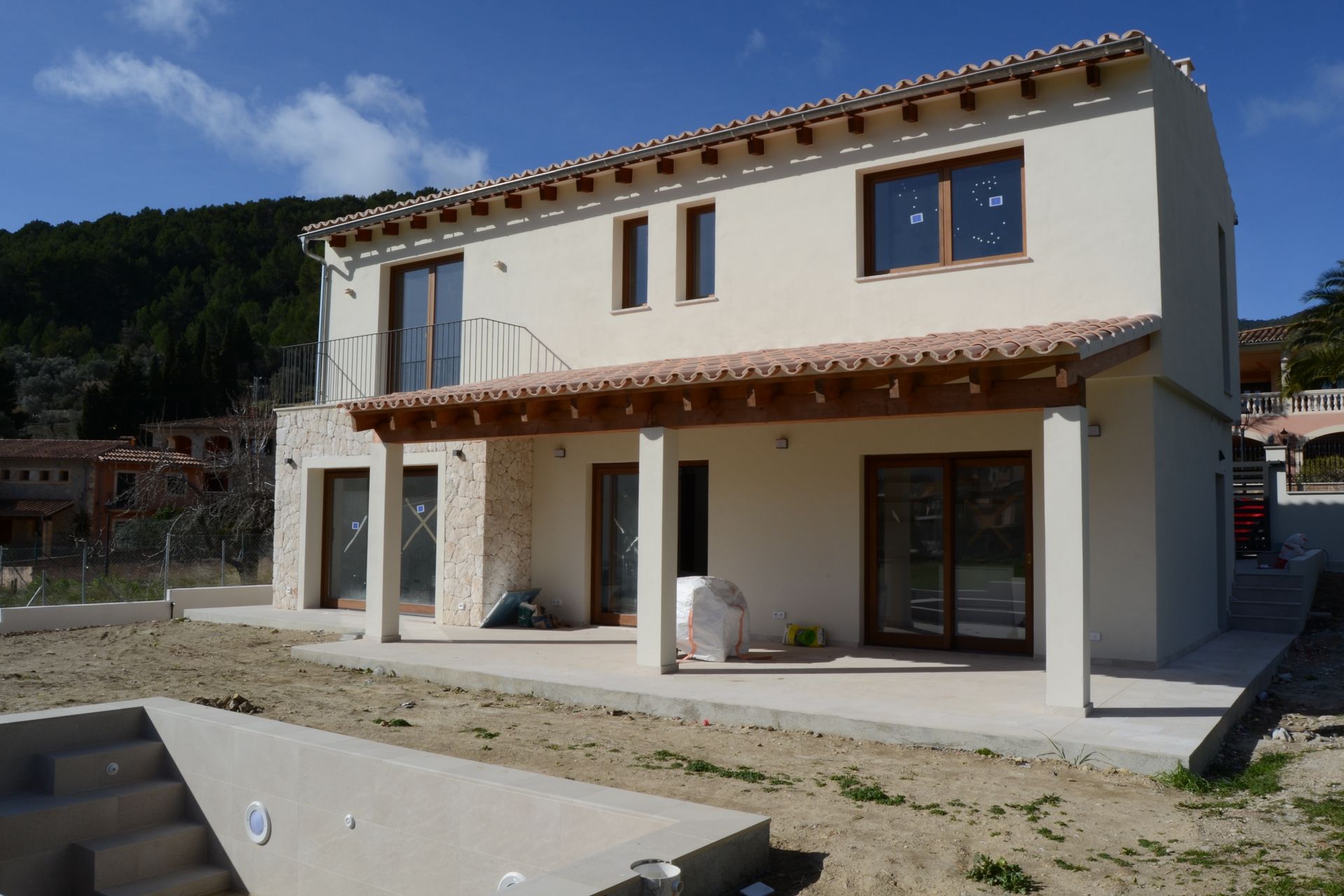 Casa de dos plantas color crema con tejado de terracota, porche cubierto y patio sin terminar en un entorno rural.