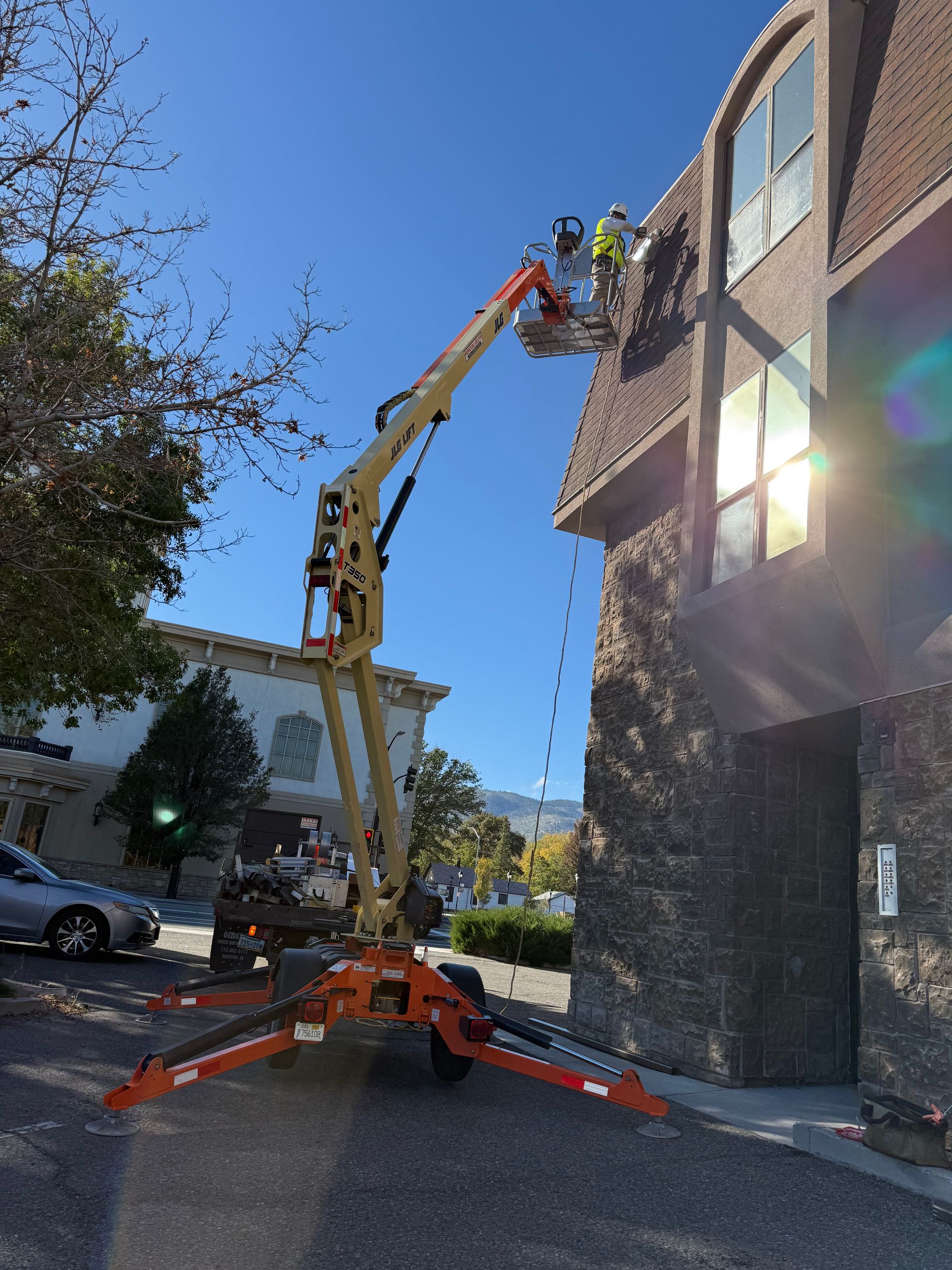Man in lift working on building exterior. Orange lift arm, blue sky, car, and trees.