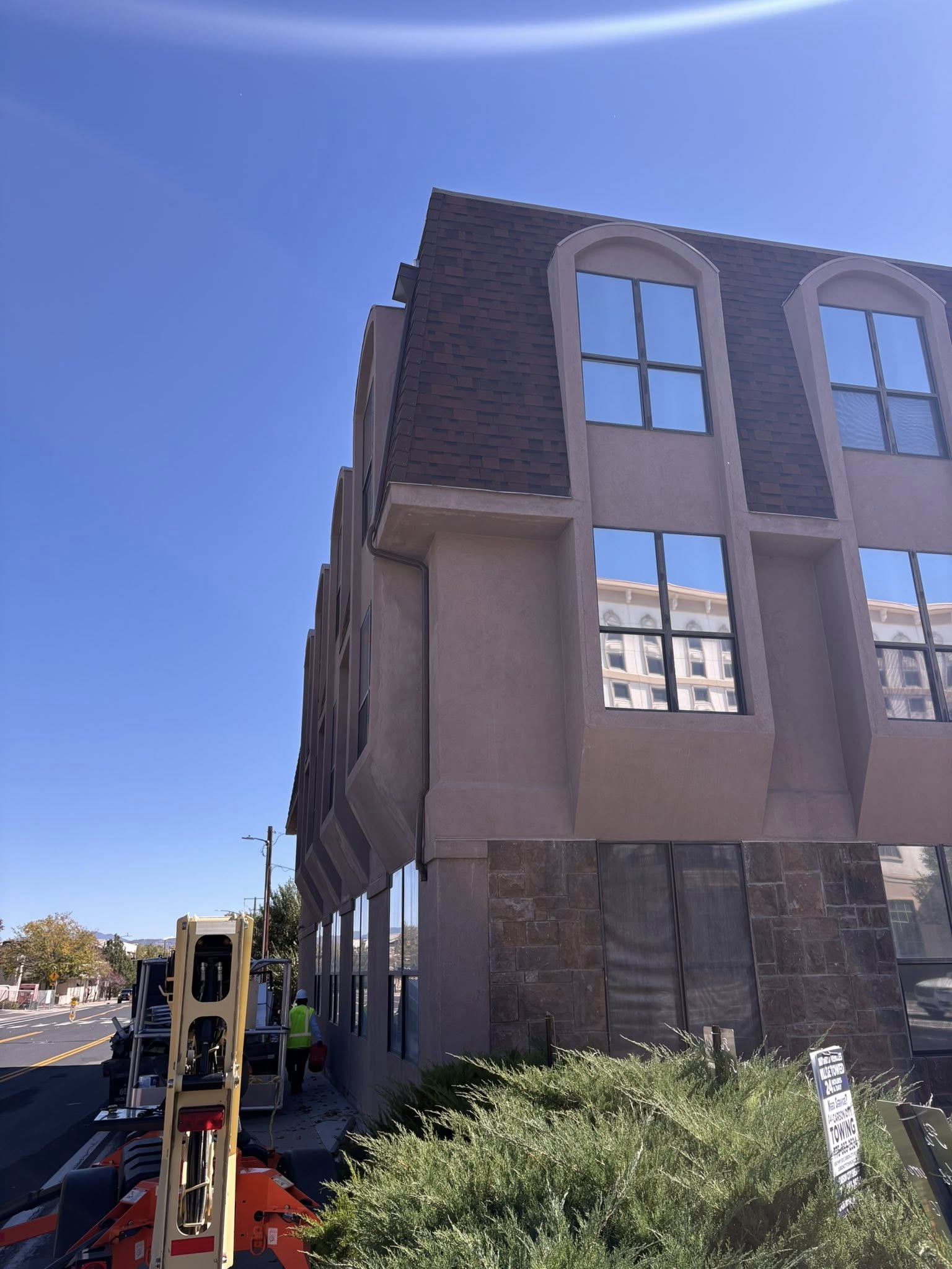 Two-story building with brown roof and mirrored windows; construction equipment and workers below.