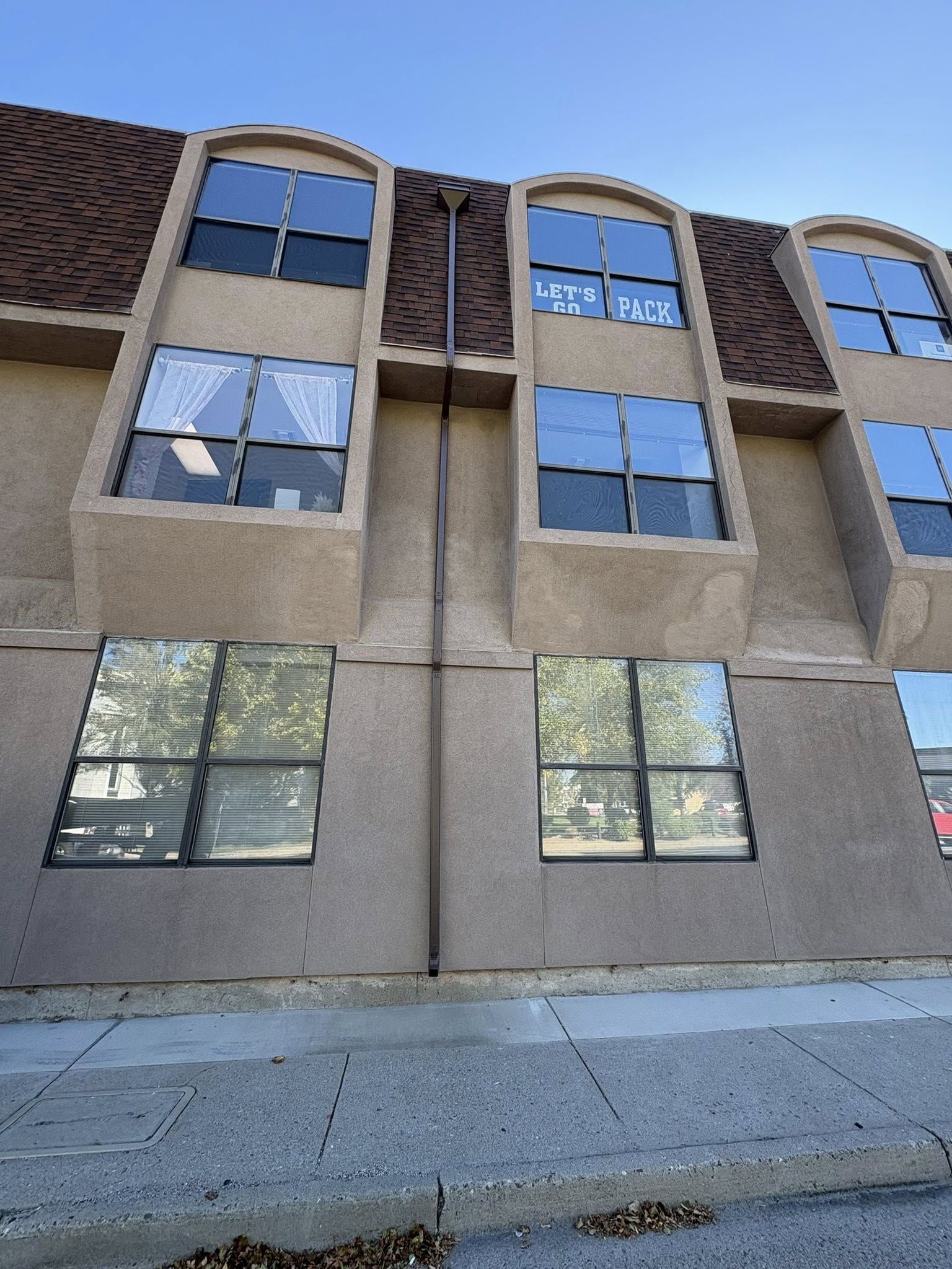 Building with multiple windows, concrete exterior, brown roof, and a sidewalk in front.